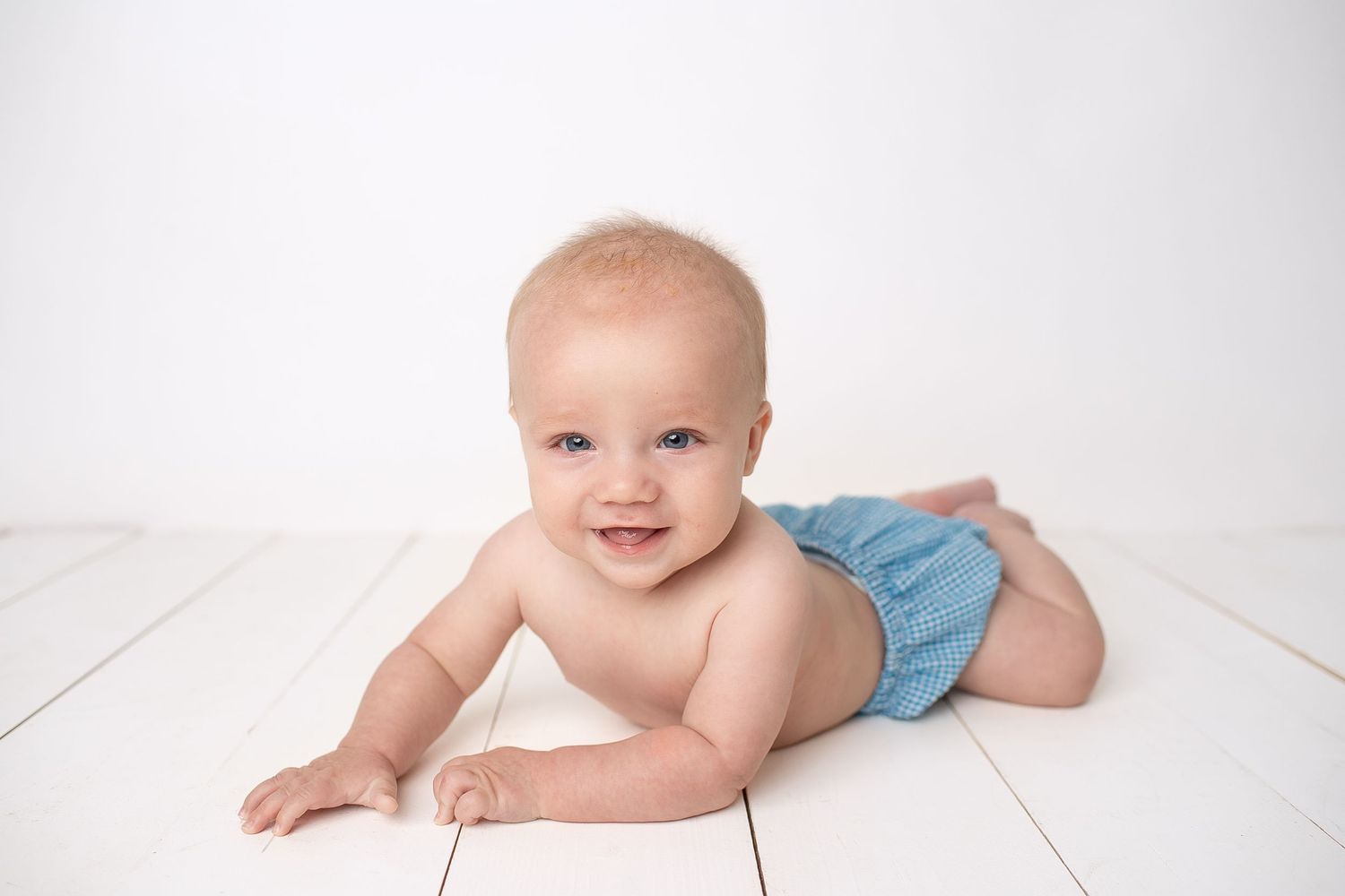Smiling baby in blue diaper lying on white wooden floor against white background.
