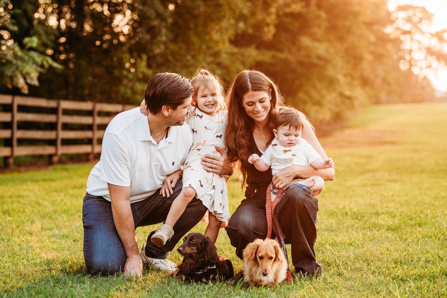 Happy family with two young children and two dogs enjoying golden hour outdoors on lush green grass near a wooden fence.