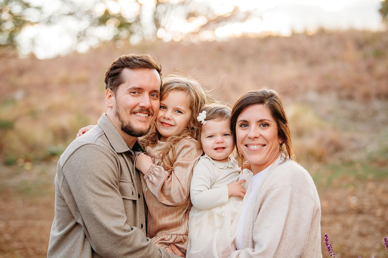 Smiling family of four posing together in a golden autumn field, parents holding two young daughters.