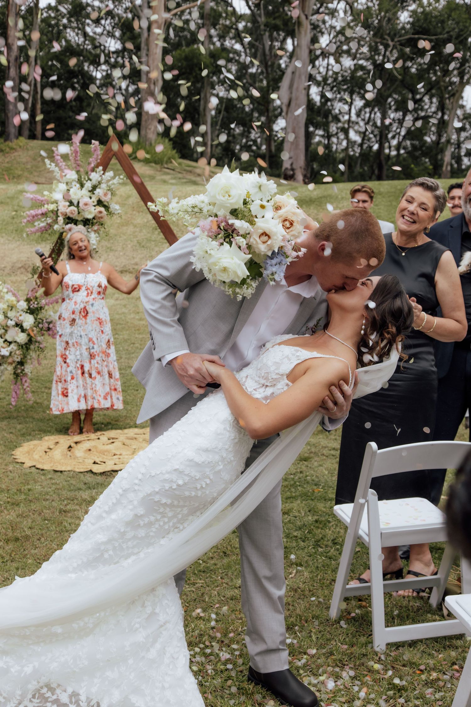 A joyful wedding celebration outdoors with a couple sharing a romantic dip kiss surrounded by flowers and guests.