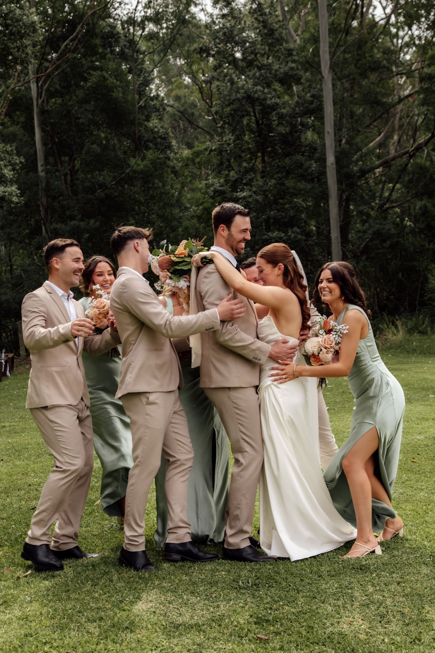 Wedding party in beige suits and sage green dresses performs a fun falling domino pose on grassy lawn.