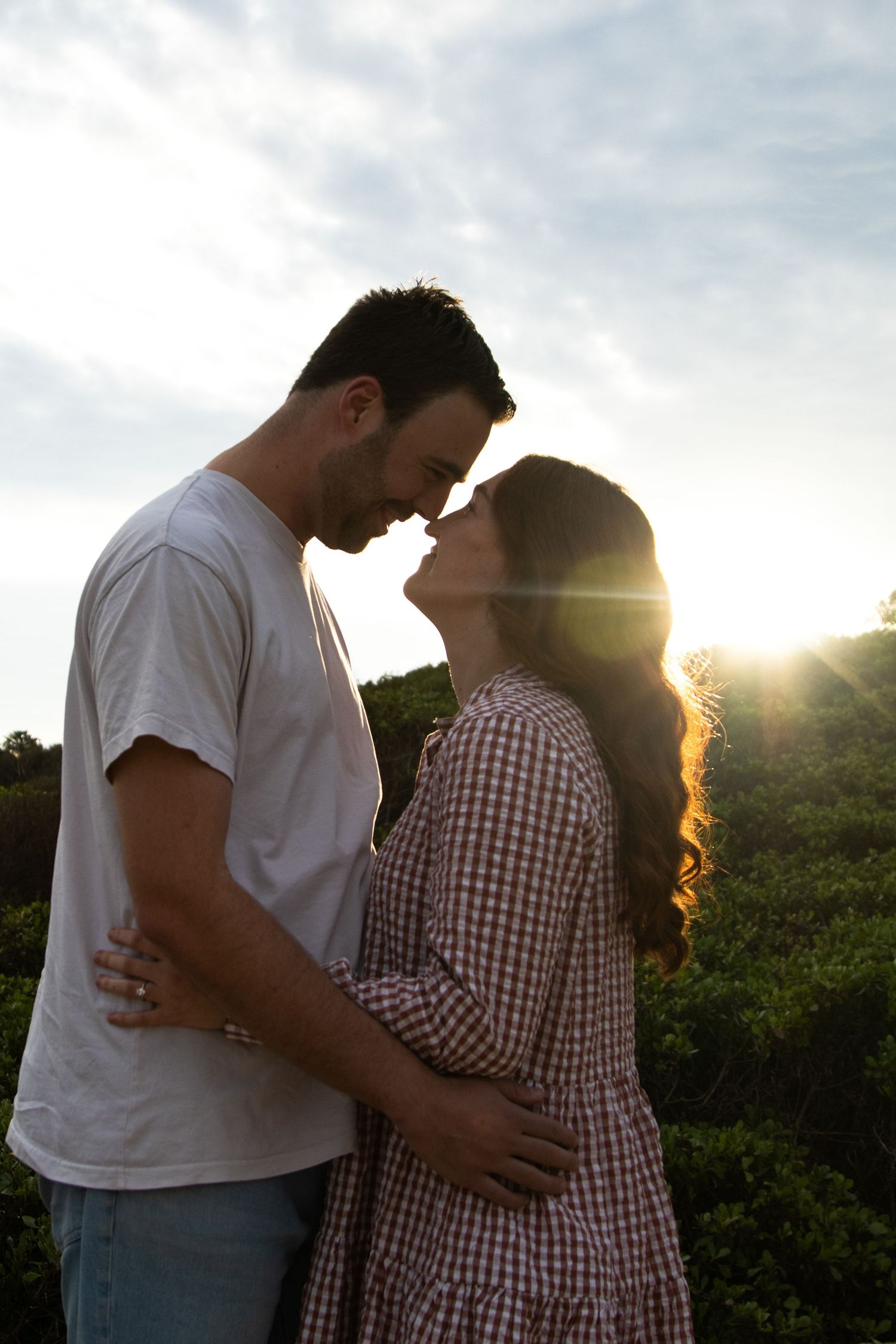 Romantic couple embraces in silhouette against sunset with sunbeams shining through trees in background.