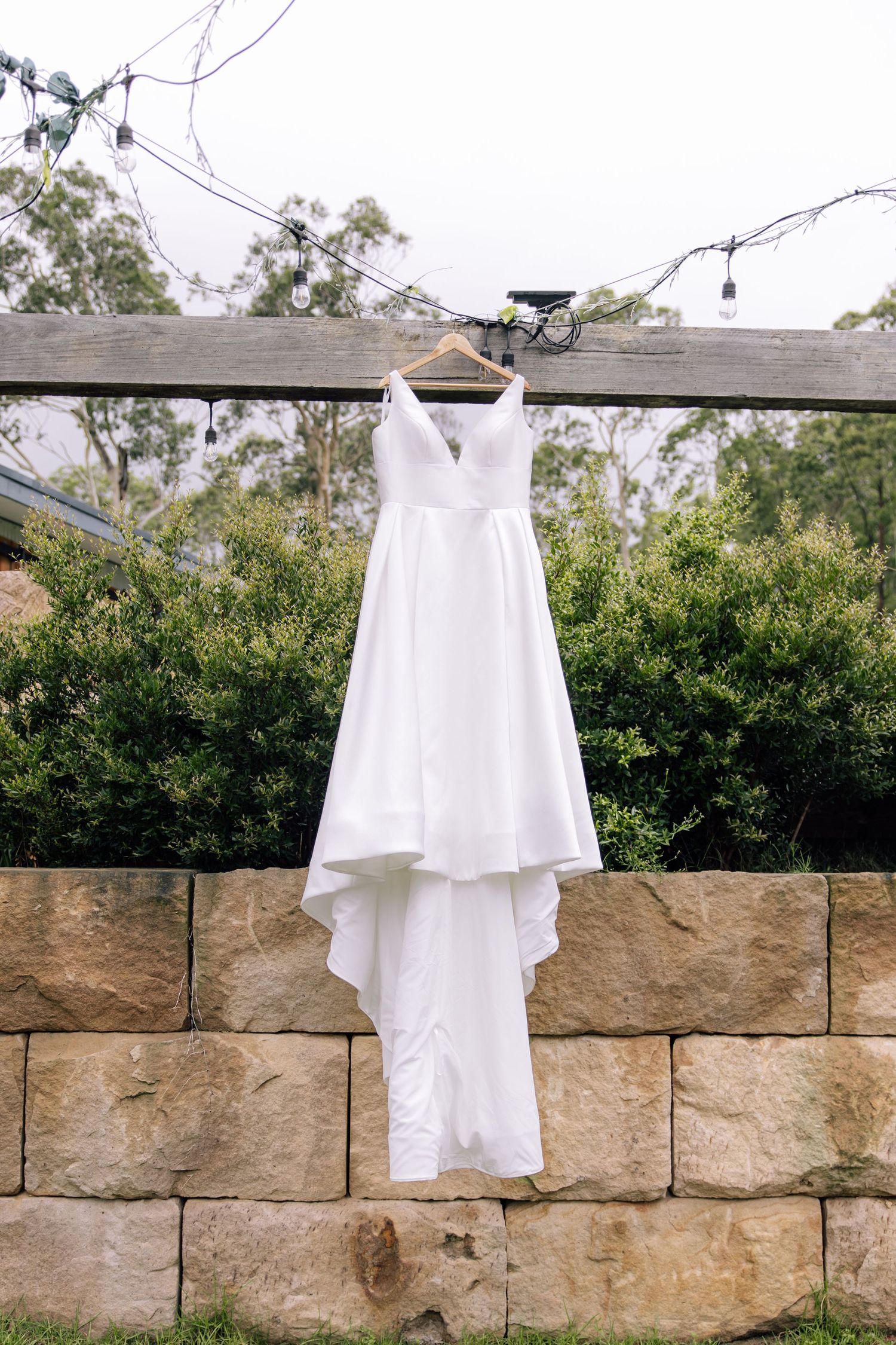 A white wedding dress hangs elegantly against a rustic stone wall backdrop in an outdoor garden setting.