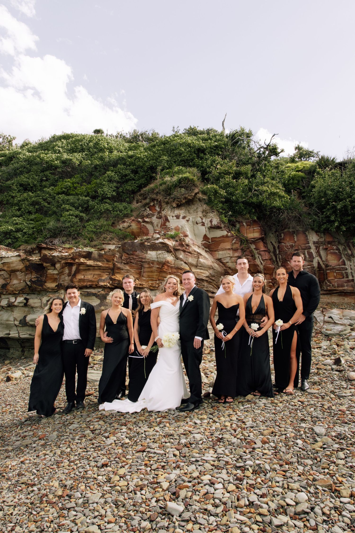 Wedding party in black dresses and white gown pose together on rocky beach against red cliffside backdrop.