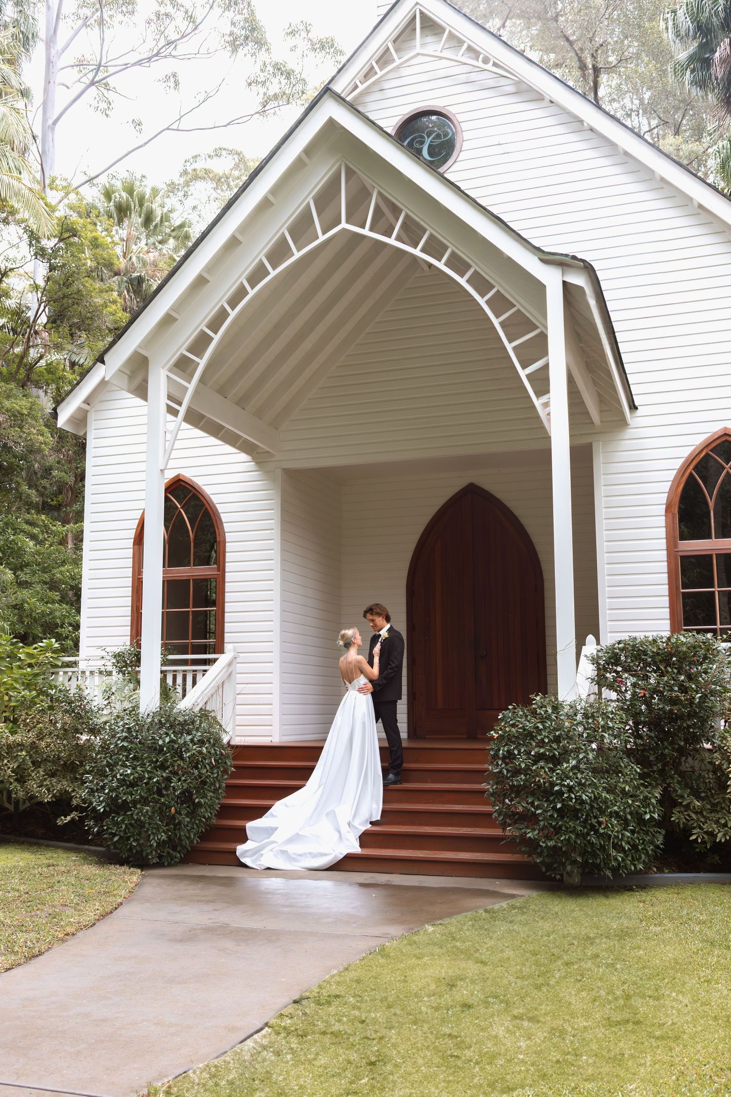 A charming white chapel with arched Gothic windows and a couple embracing on the red-brick steps at its entrance.