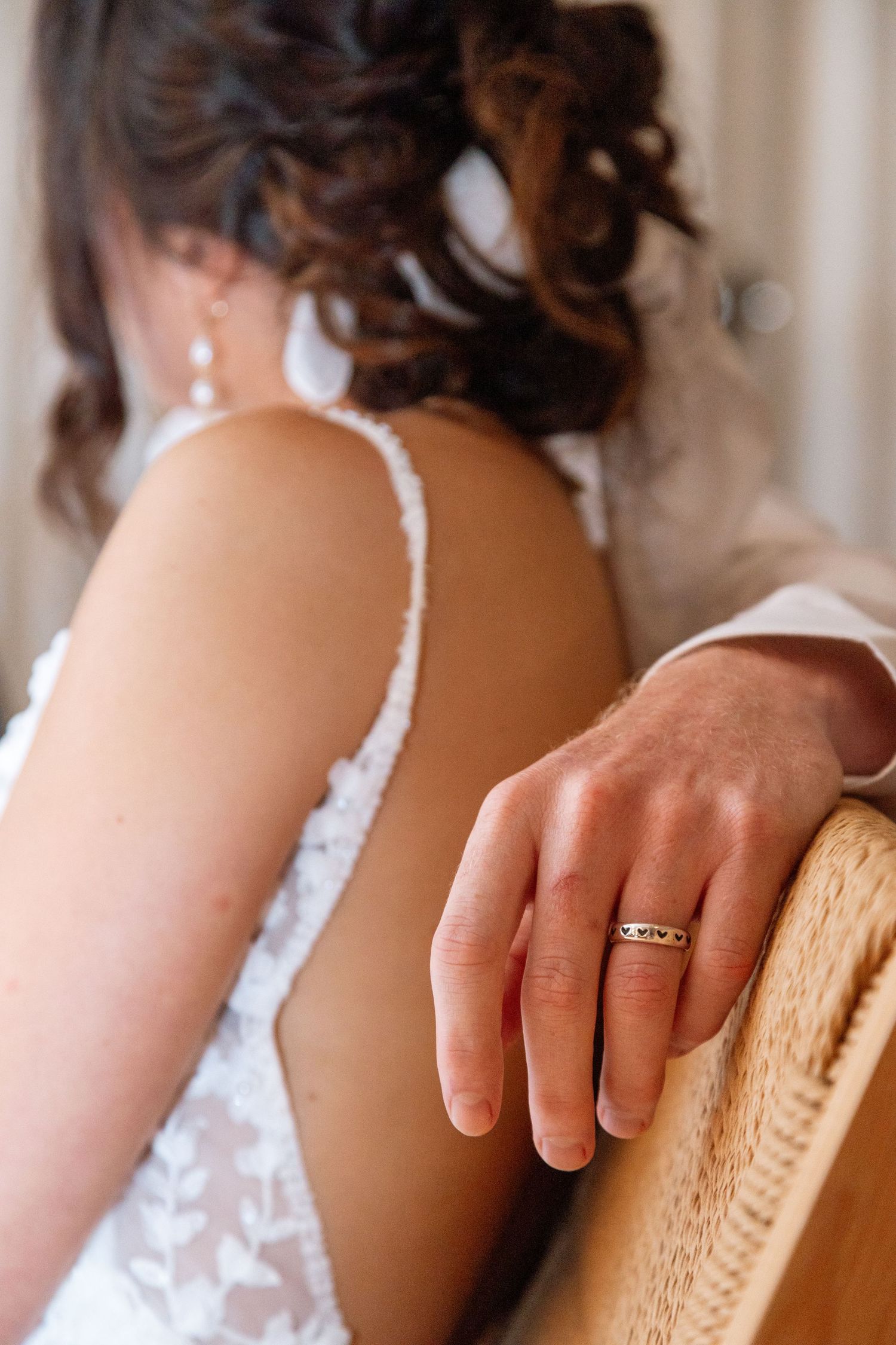 Close-up view of a delicate engagement ring worn on a hand resting against a wicker chair, with lace details visible.
