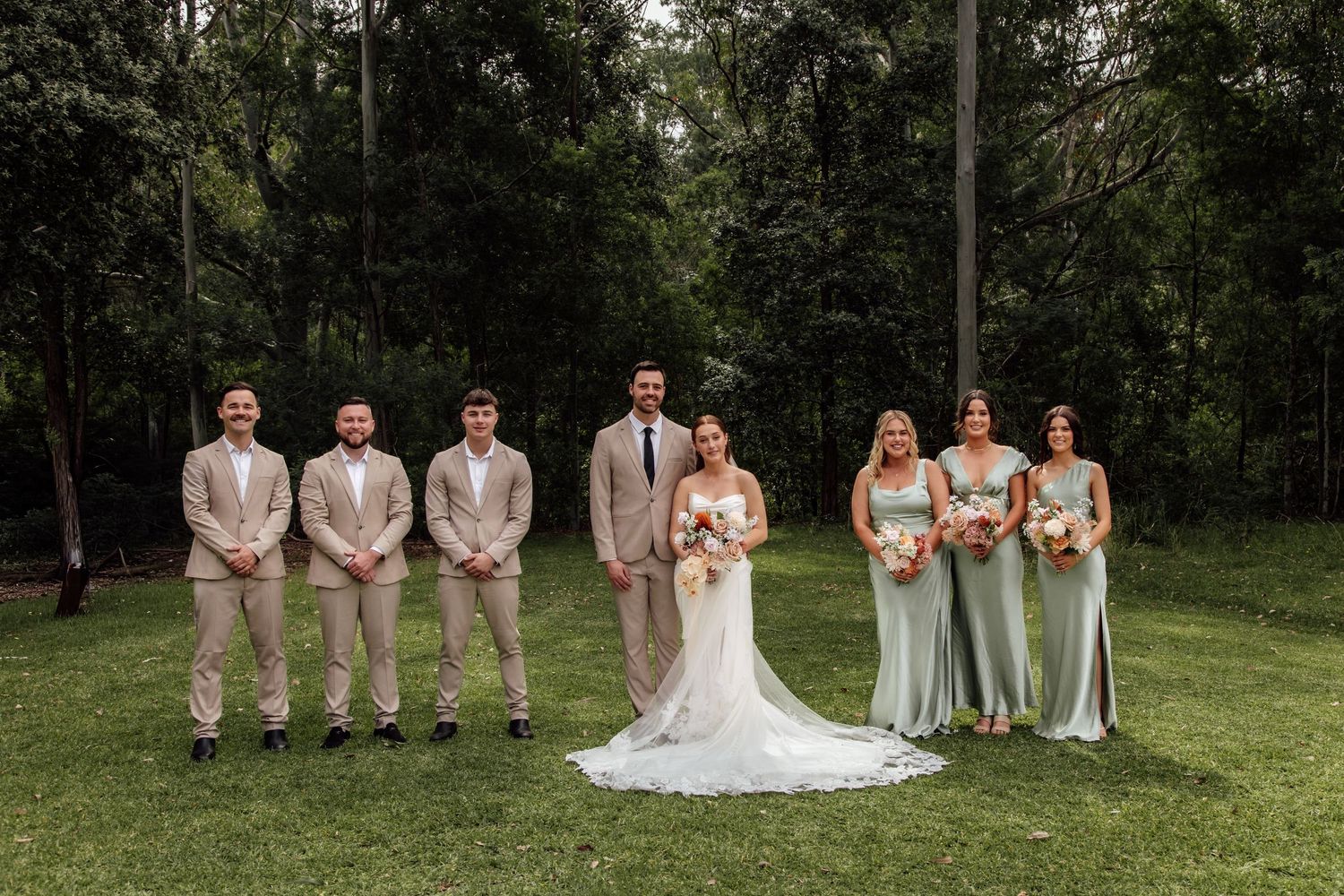 Wedding party in sage green and beige attire poses on grassy lawn with forest backdrop.