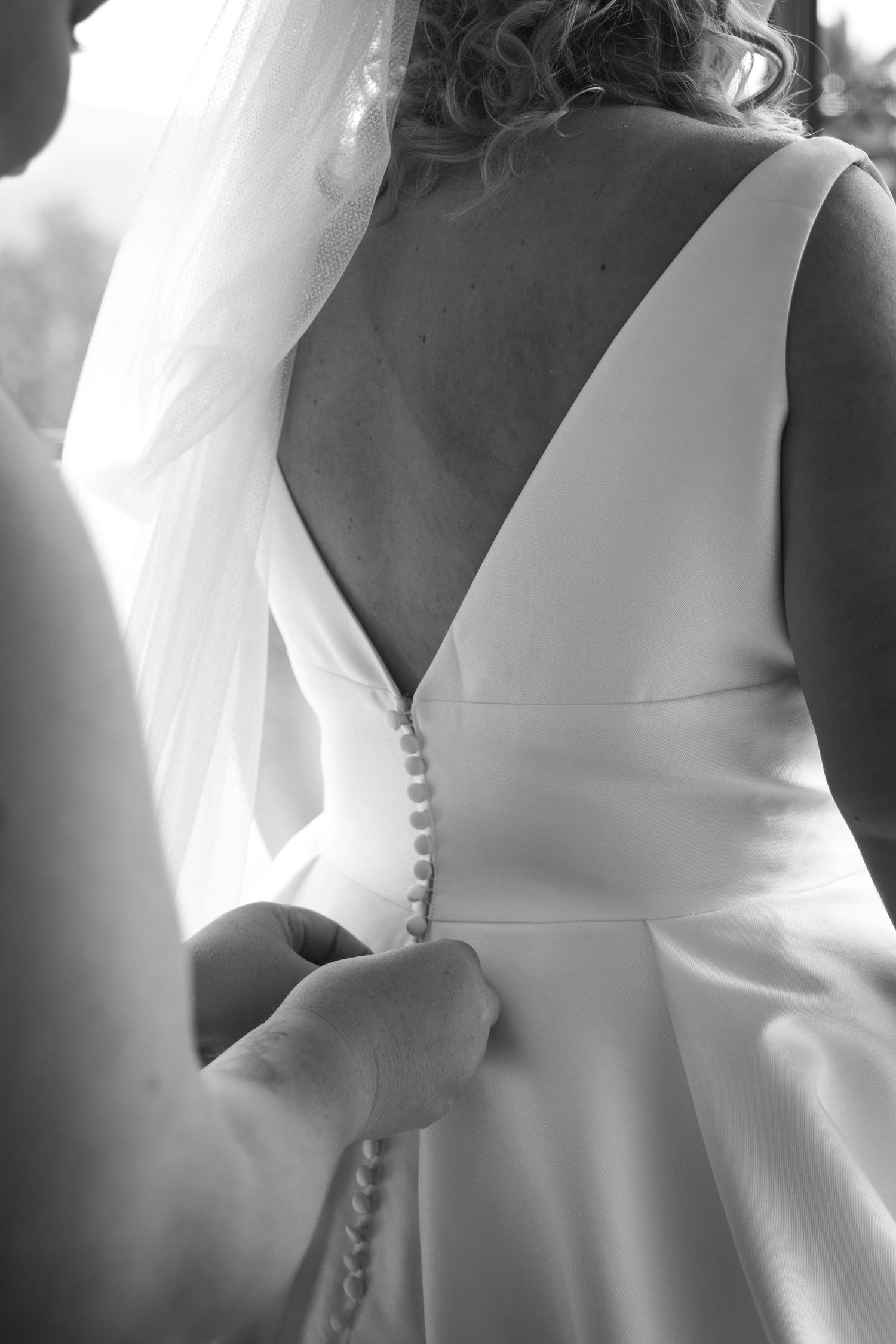 Close-up black and white photo of hands buttoning up the back of an elegant white wedding dress with covered buttons.