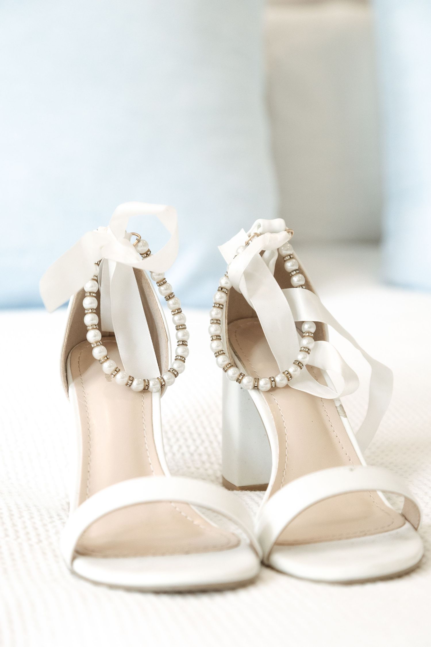 Elegant white bridal sandals with pearl ankle straps and delicate strappy design on textured fabric background.