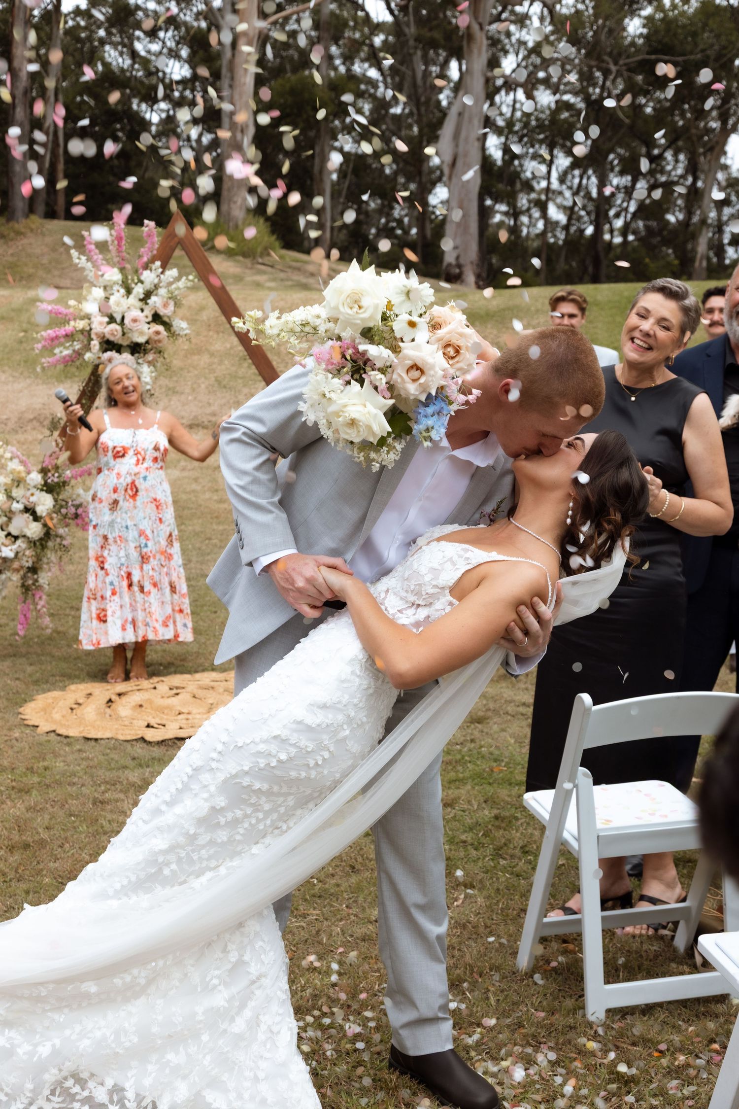 Newlyweds share romantic dip kiss at outdoor wedding ceremony while guests throw flower petals in celebration.