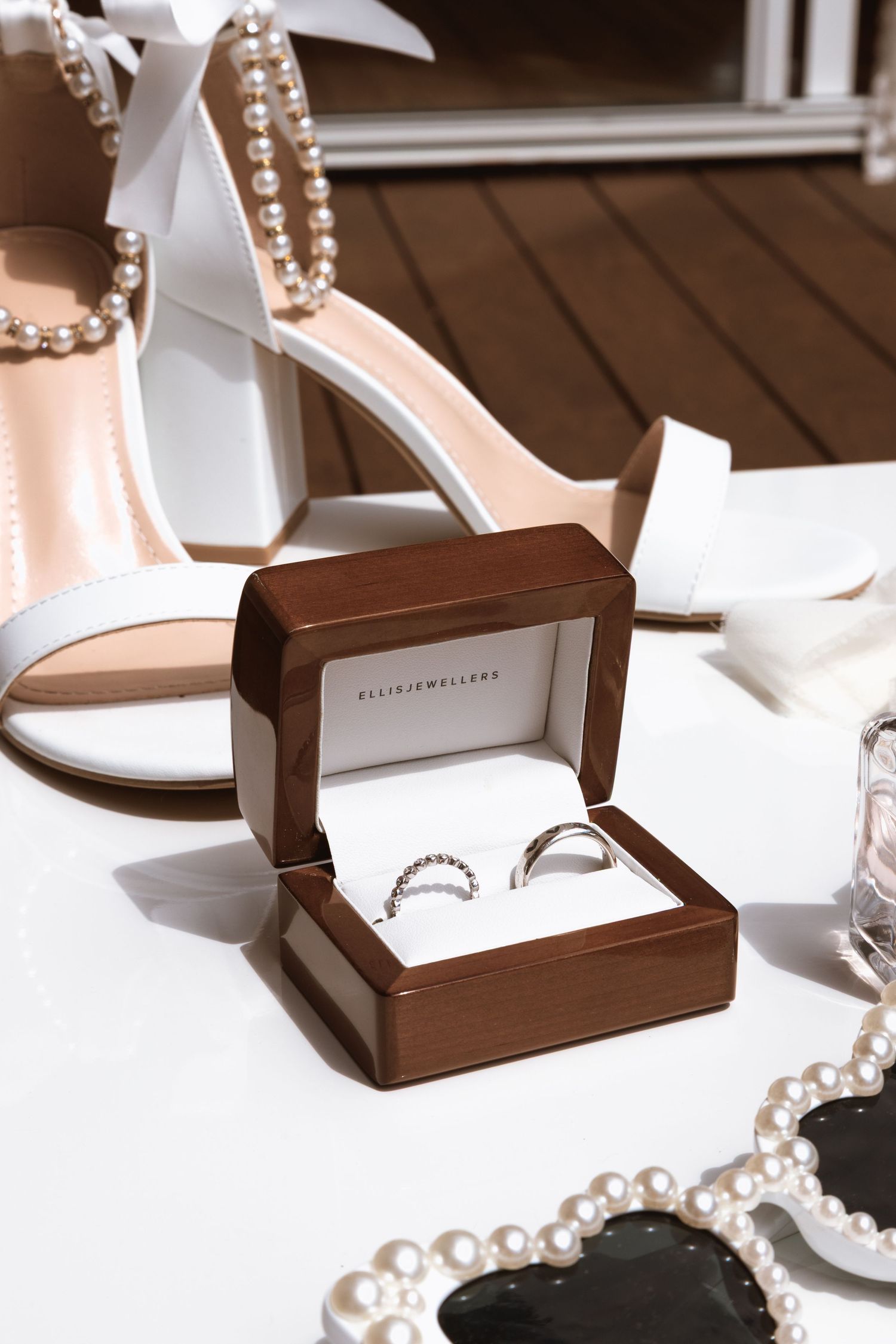 Wedding rings in a brown wooden box displayed with white heels and pearl accessories on a wooden surface.