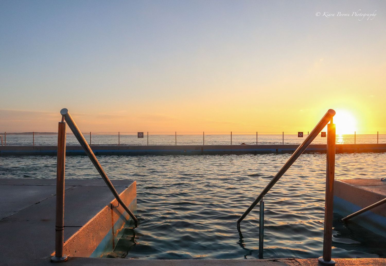 Handrails leading into an ocean pool at sunset with golden sun reflecting off rippling water.