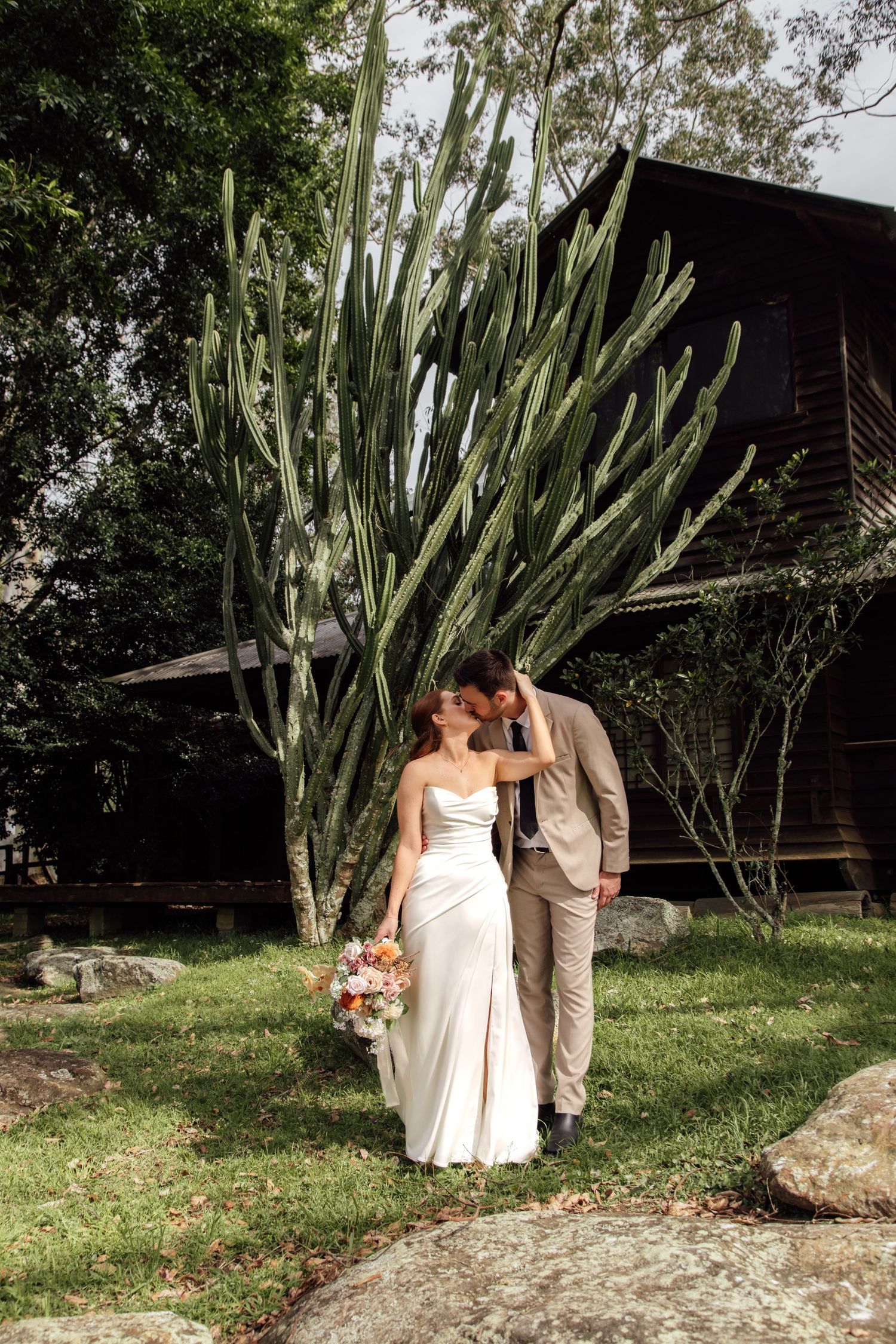 A couple in wedding attire poses together in a garden setting with tall cactus plants in the background.
