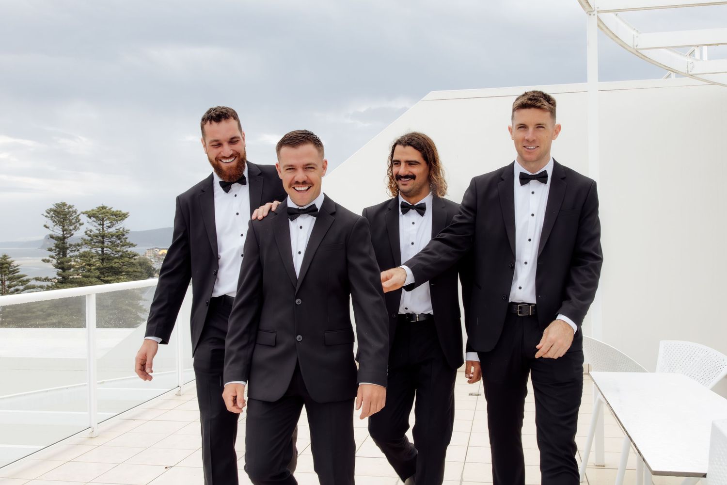 Four groomsmen in black tuxedos and bow ties walk together smiling on a rooftop terrace.