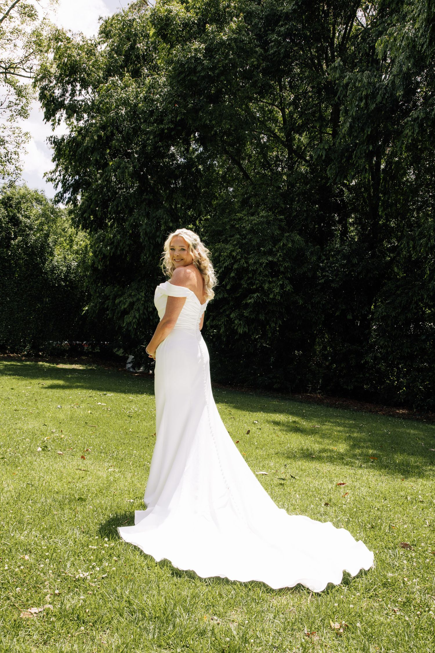 A bride in a flowing white wedding dress poses gracefully on a sunlit lawn surrounded by lush green trees.