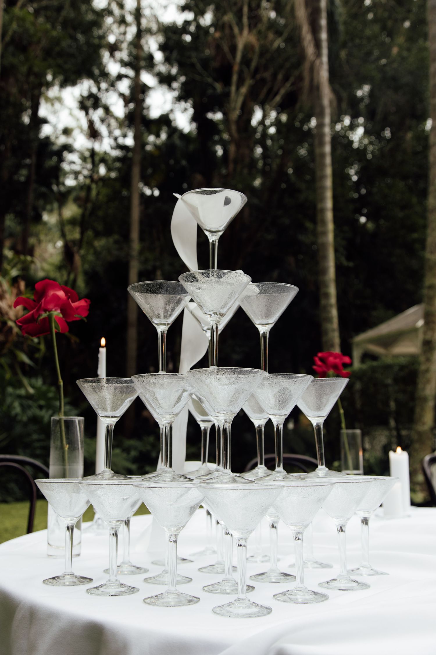 Empty martini glasses arranged in rows on white tablecloth with red flowers in outdoor garden setting.