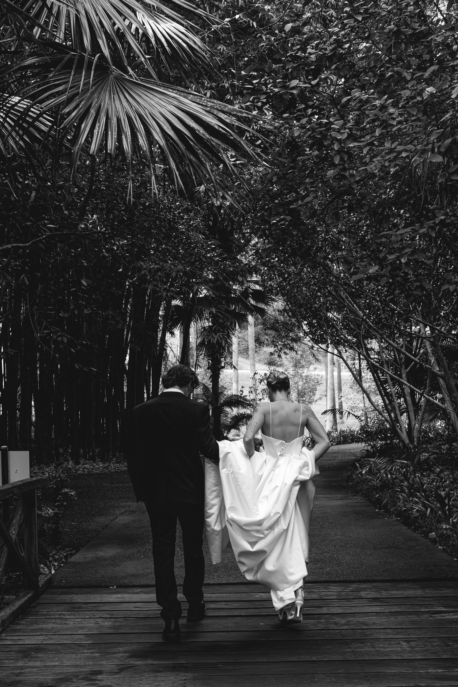 Black and white photo of a newlywed couple walking away on a wooden path lined with tropical palm trees and foliage.