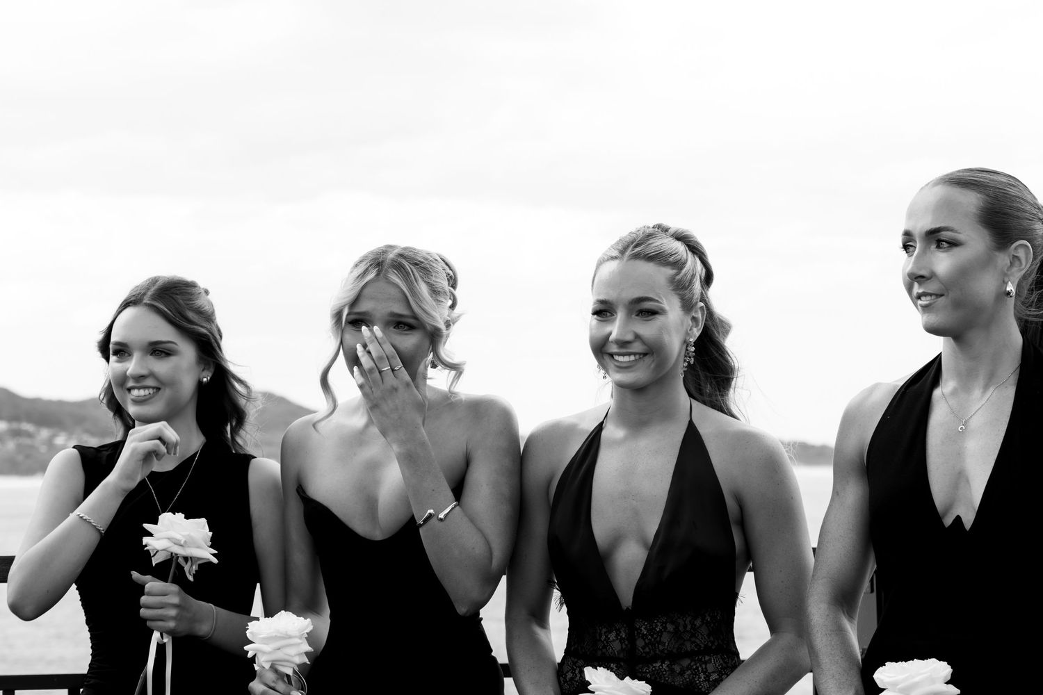 Four bridesmaids in black dresses share a candid laughing moment together at an outdoor wedding.