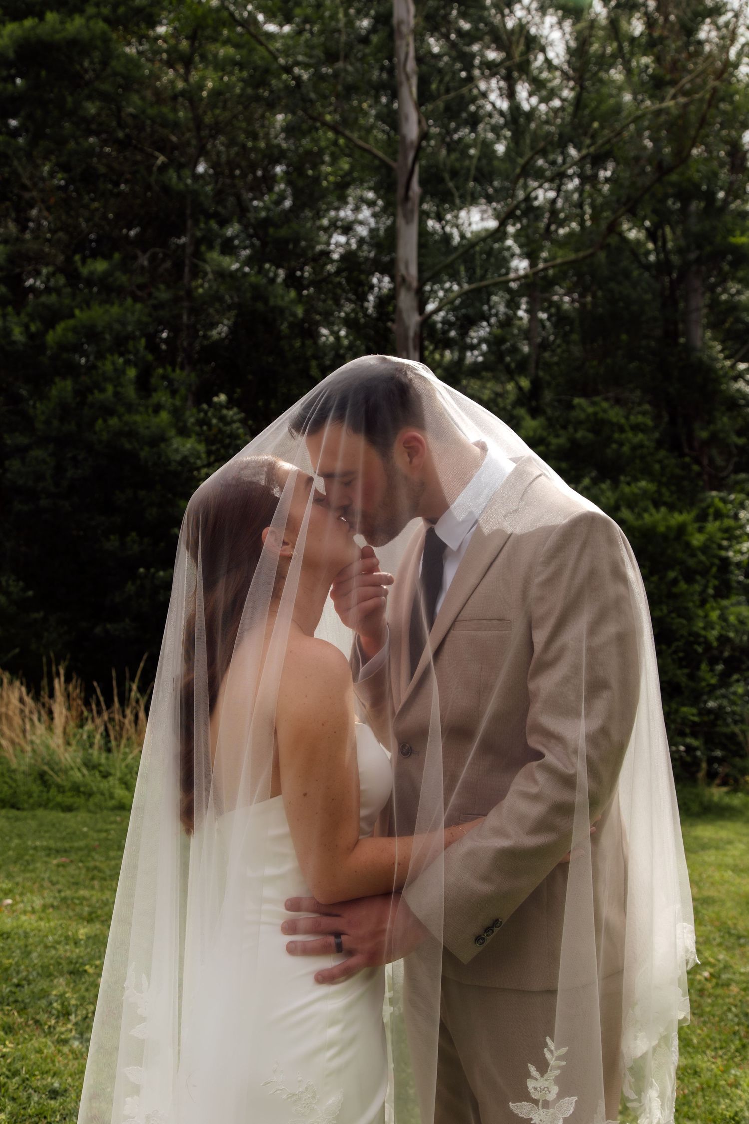 A romantic wedding moment captured outdoors with a couple embracing under a flowing white veil against a natural backdrop.