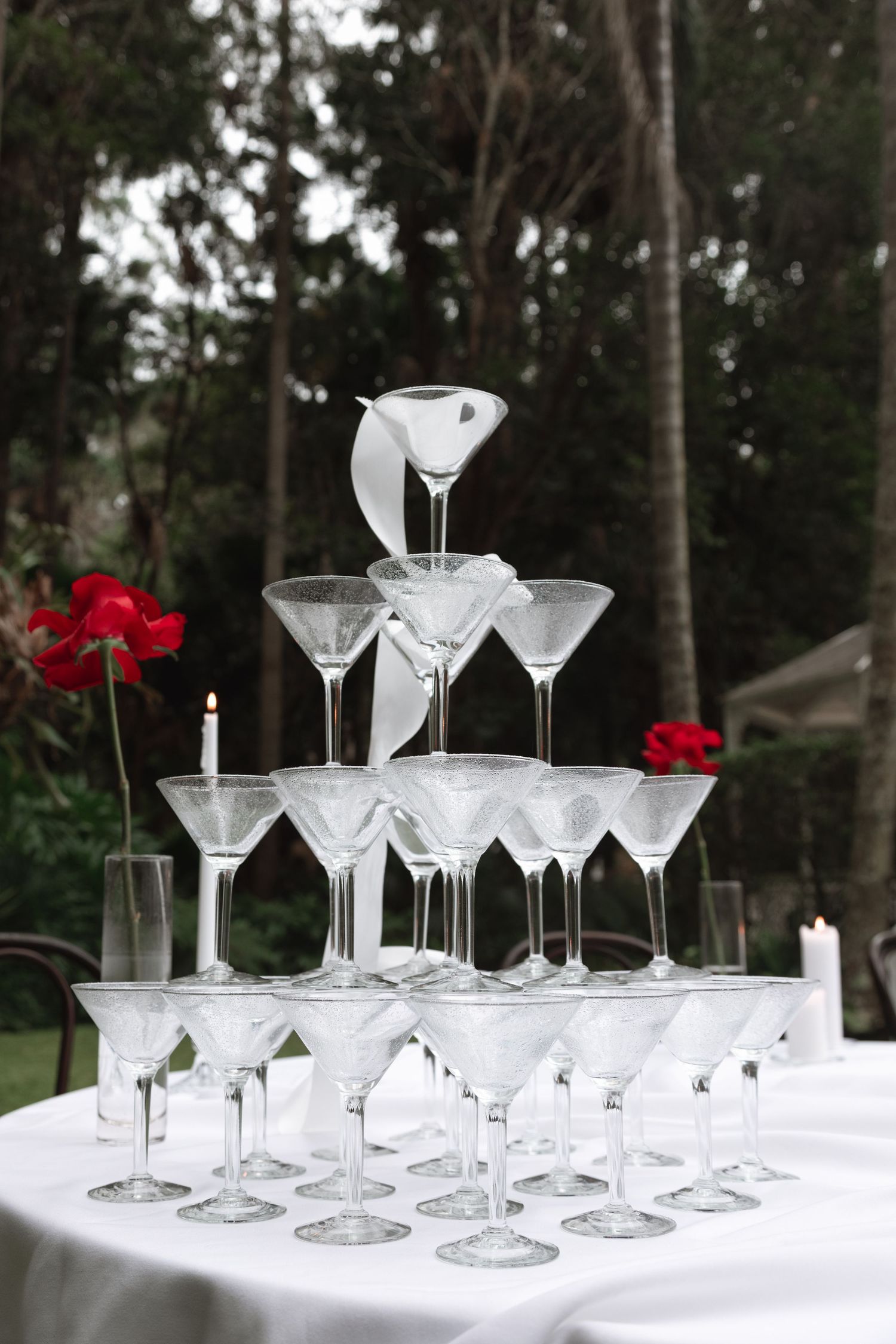 Four elegant martini glass displays arranged on white tablecloths with red floral accents and trees in the background.