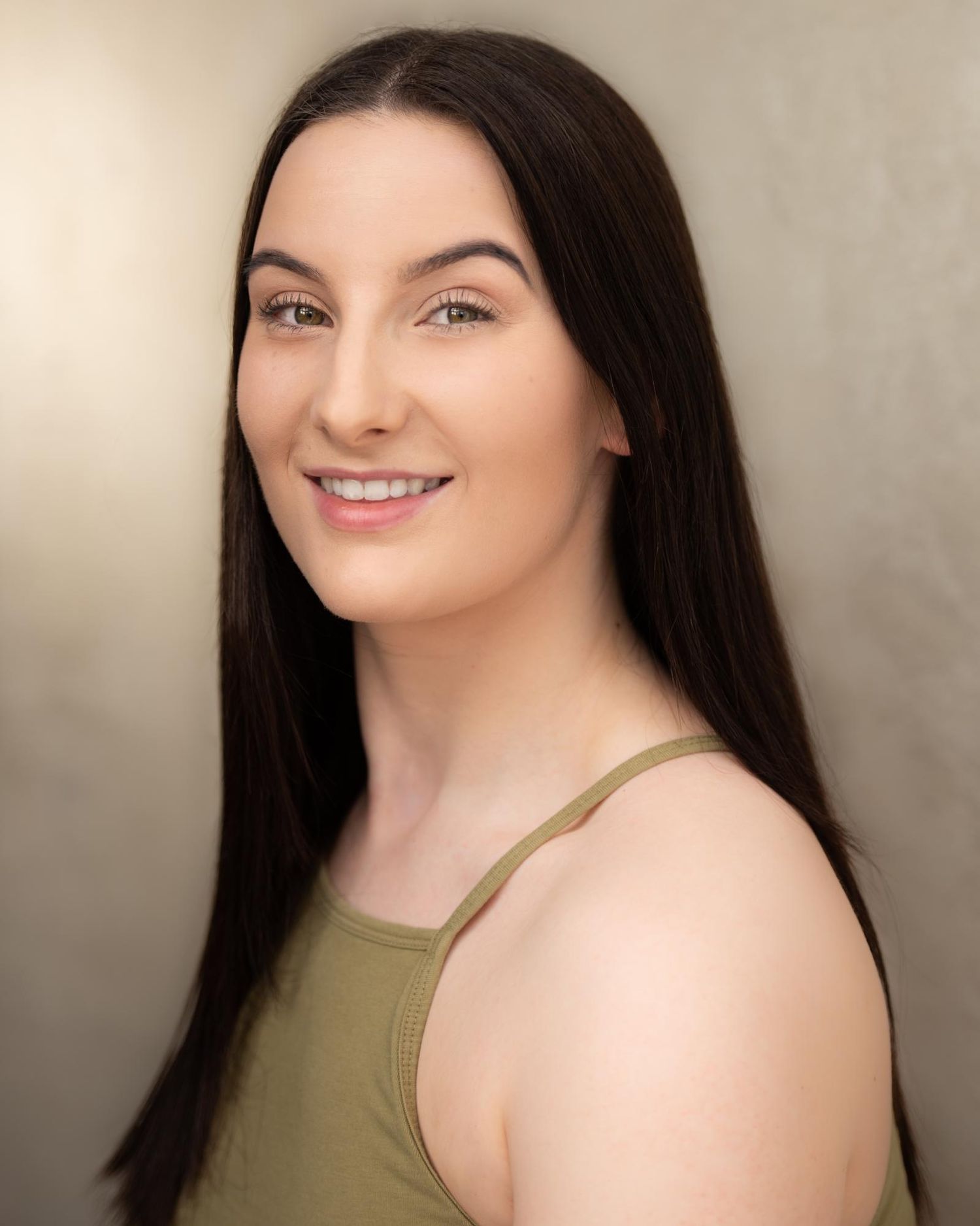 Portrait of someone with long dark hair wearing an olive green tank top against a neutral background, smiling warmly.