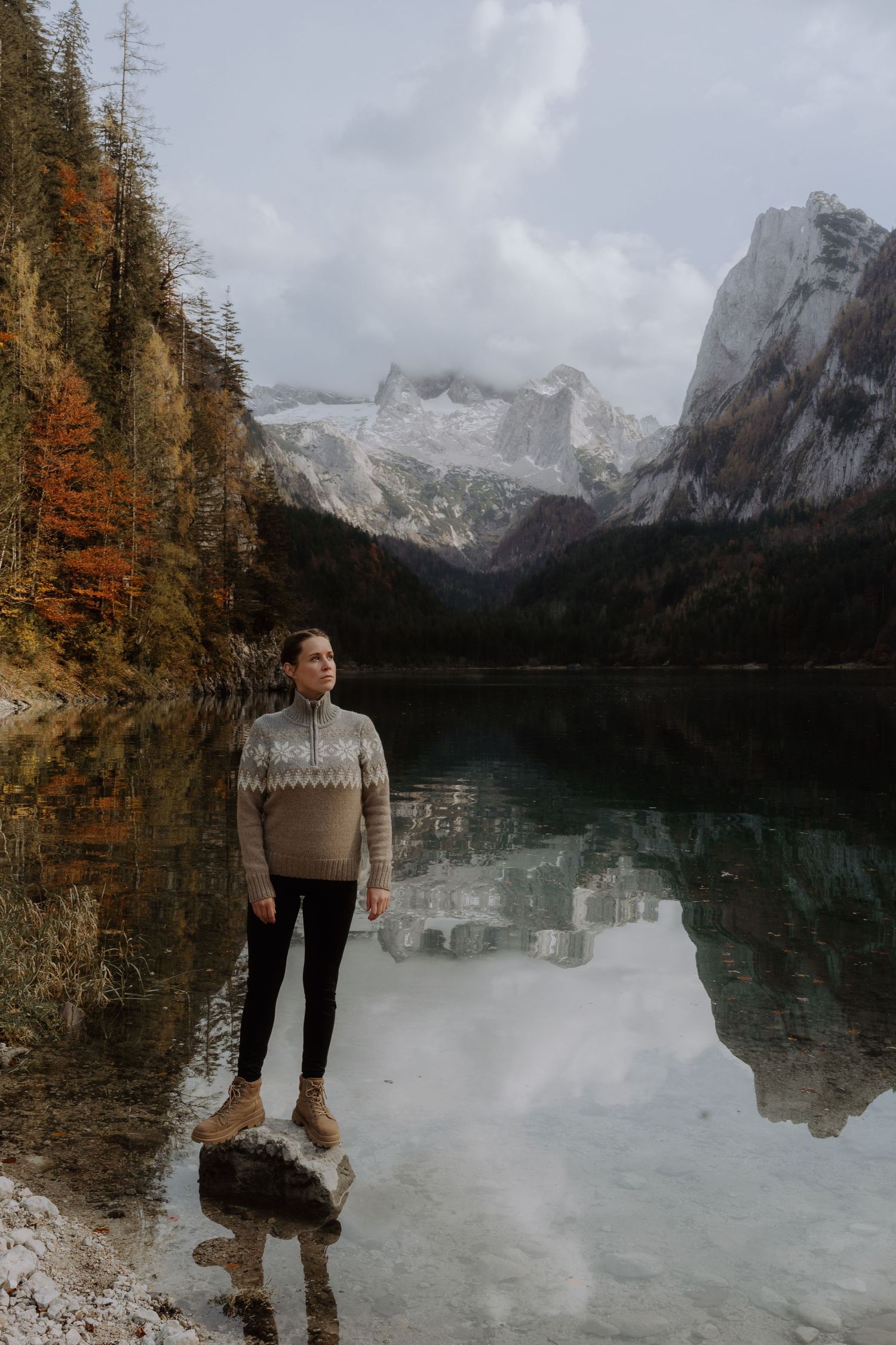 A person standing by an alpine lake with snow-capped mountains and fall foliage reflecting in water.