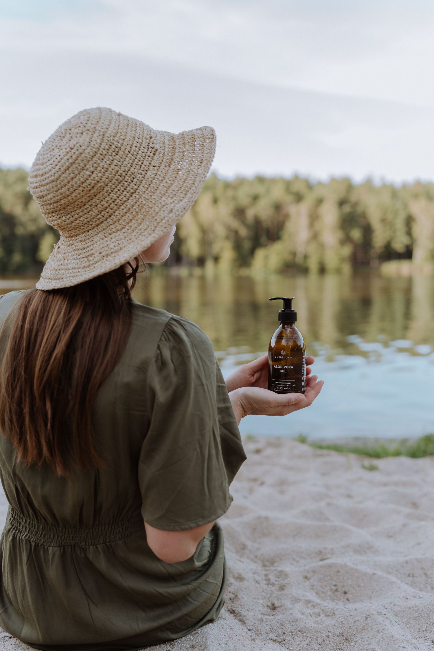 Person wearing straw hat and olive dress sitting by lake holding bottle of natural product.