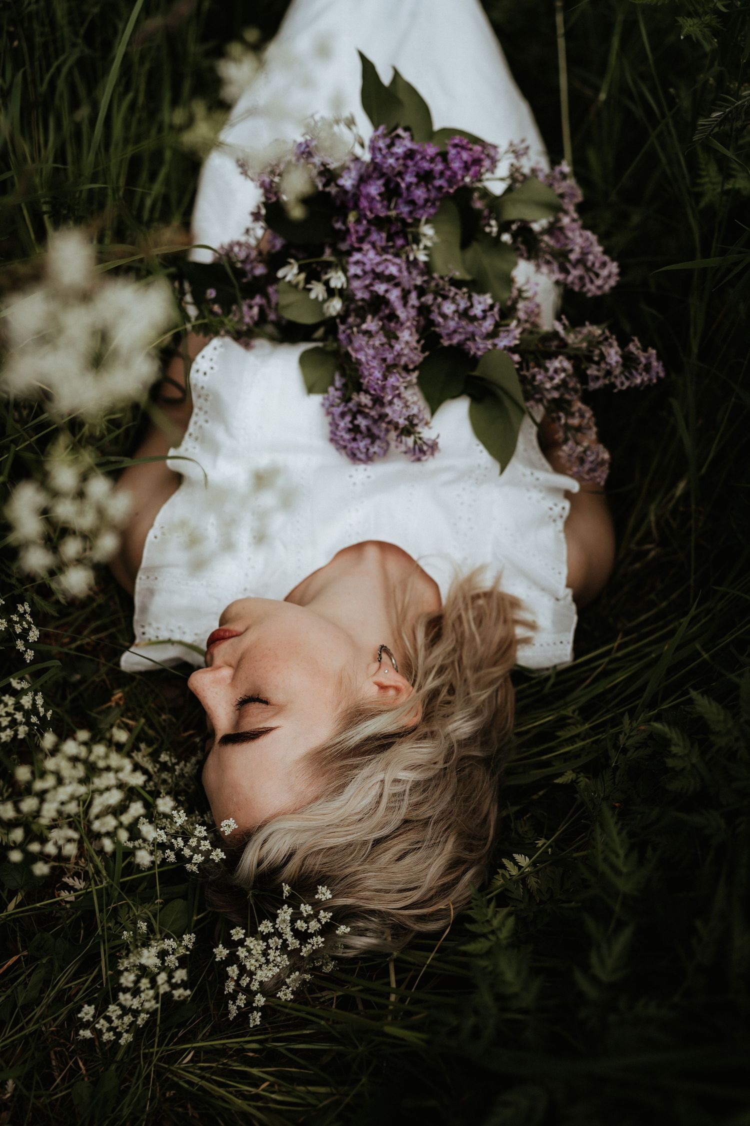 Artistic overhead shot of a woman in white lying in grass with purple lilac flowers.