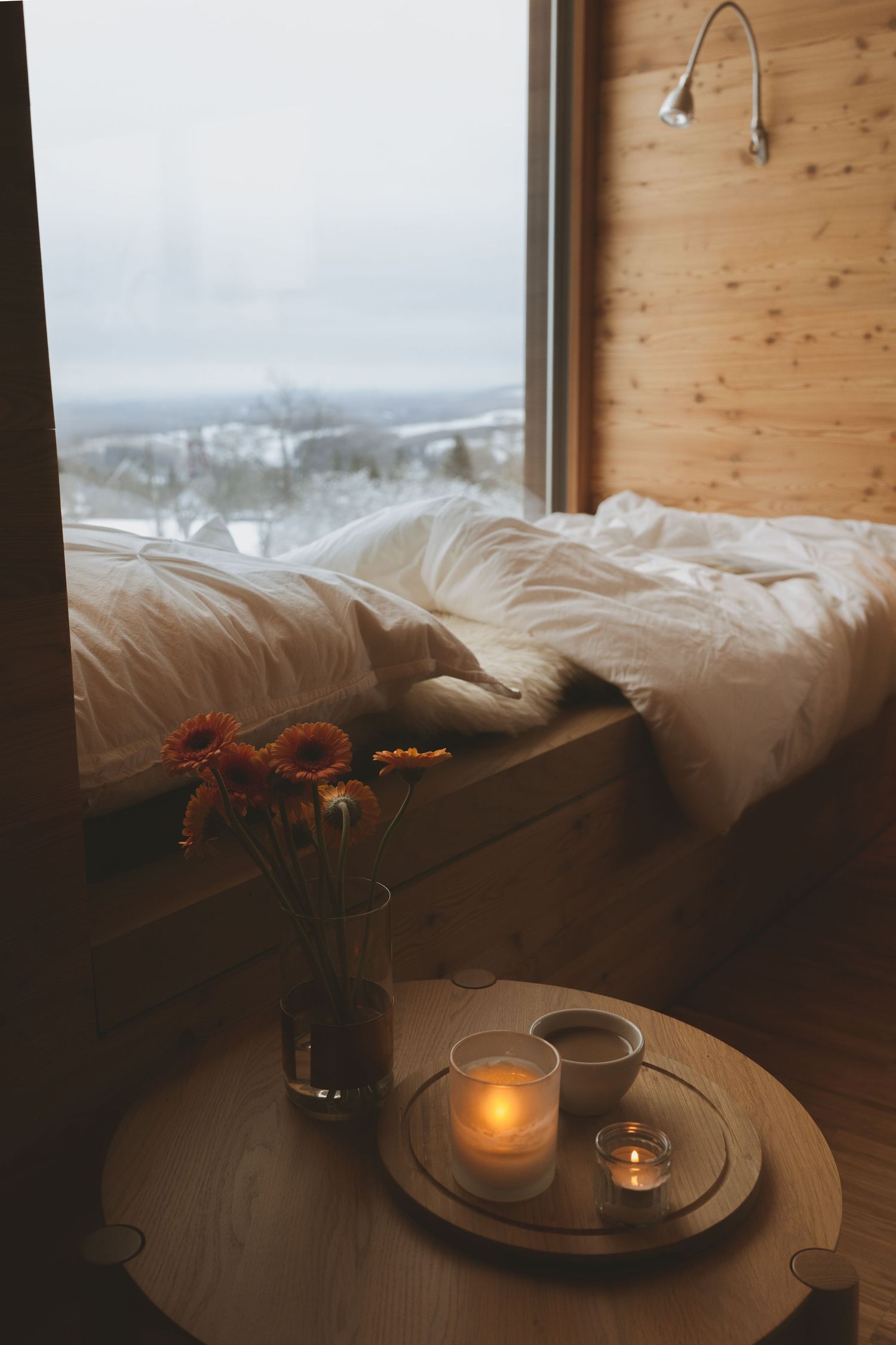 Cozy bedside scene with candles and flowers overlooking a winter landscape through wooden cabin windows.