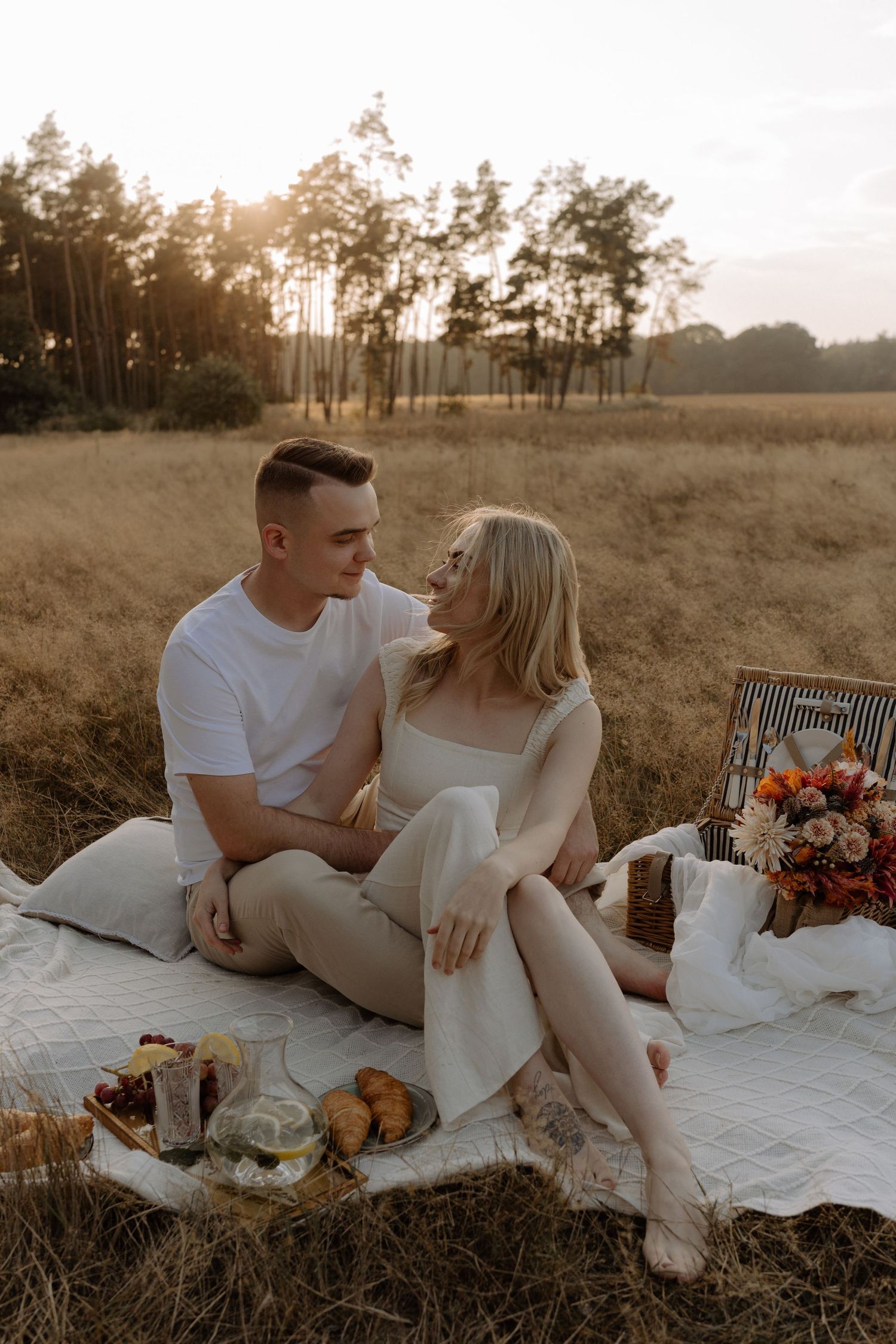 Romantic picnic scene at sunset with couple on blanket surrounded by food and flowers.