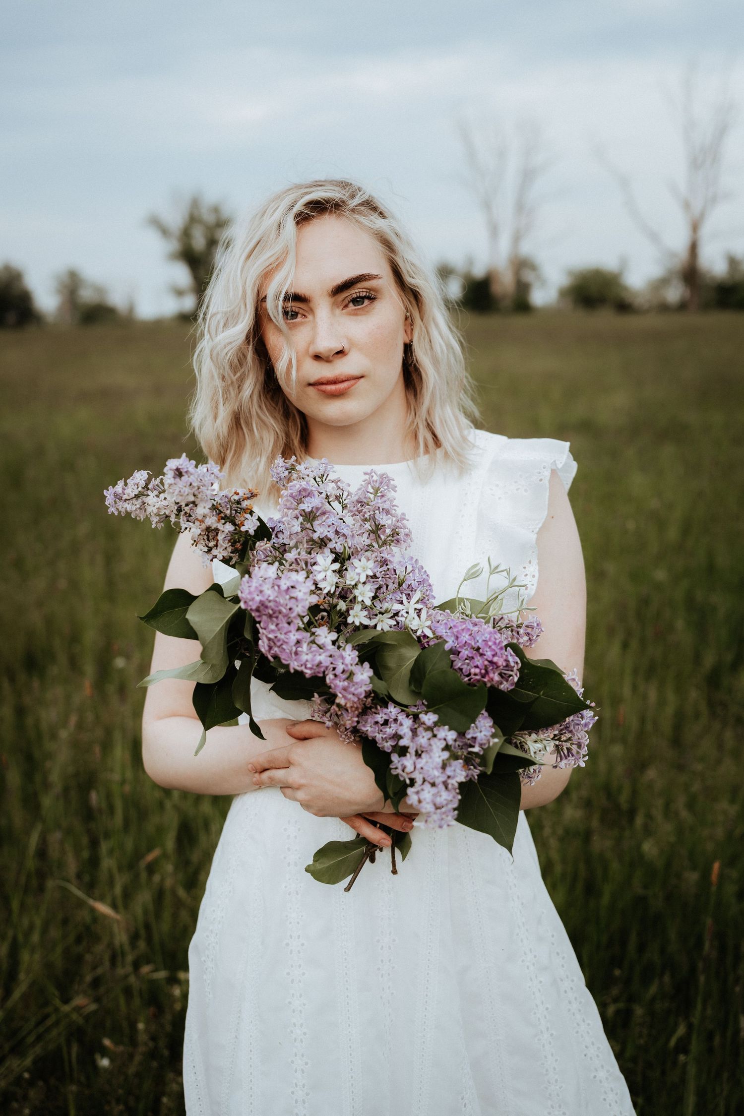 A woman in a white dress holding a bouquet of purple lilac flowers in a spring meadow.