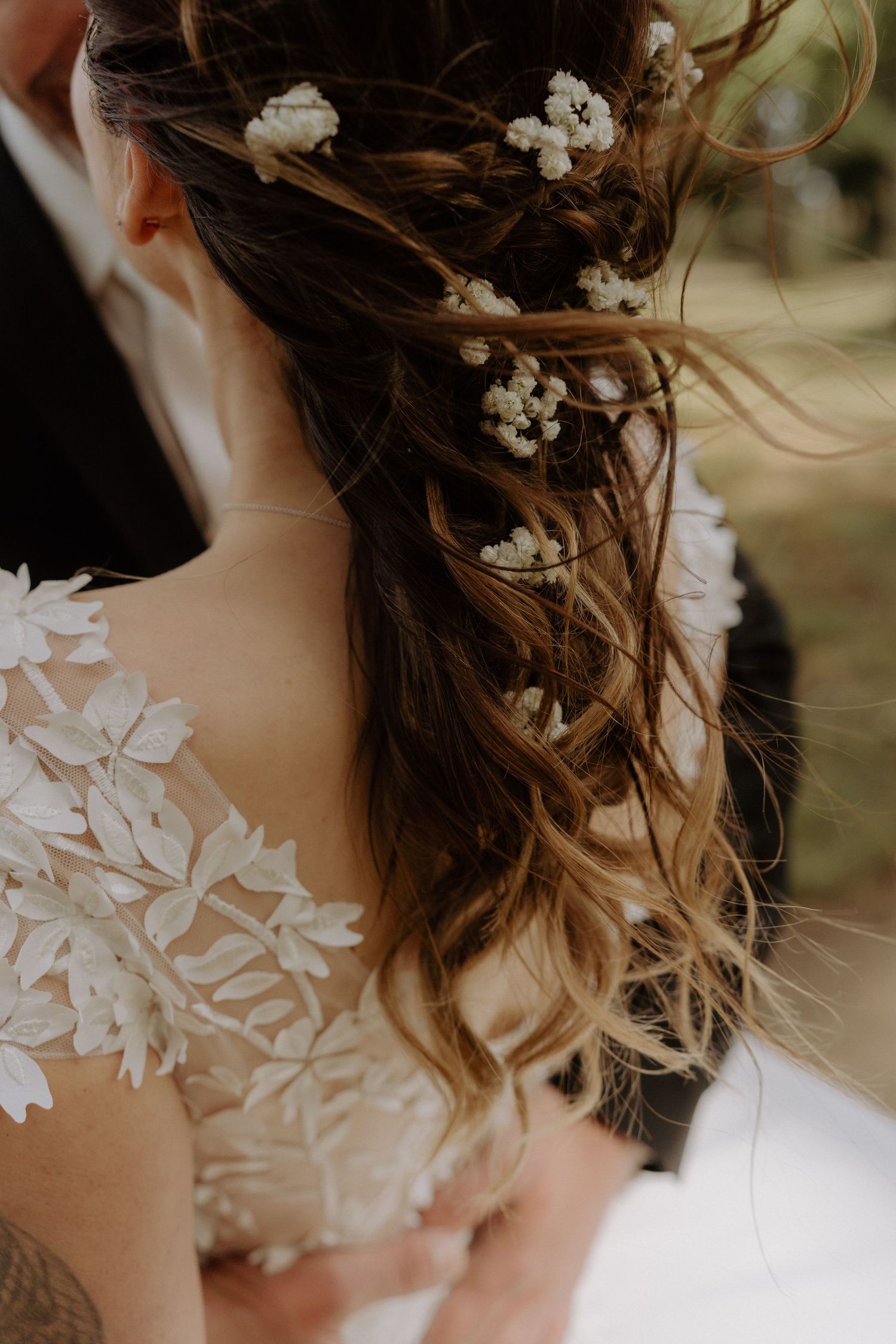 Delicate white flowers woven through loose wedding hairstyle with lace dress detail visible.