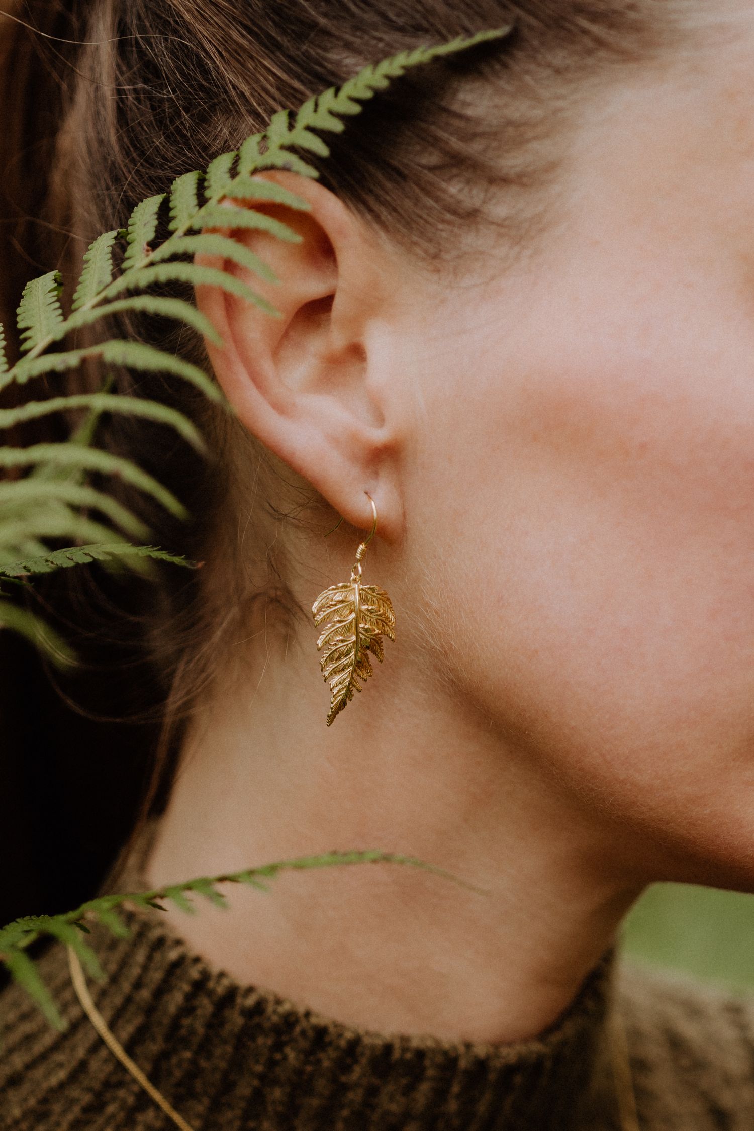 Gold leaf-shaped earring worn with fern leaves in natural lighting creating an ethereal nature-inspired composition.
