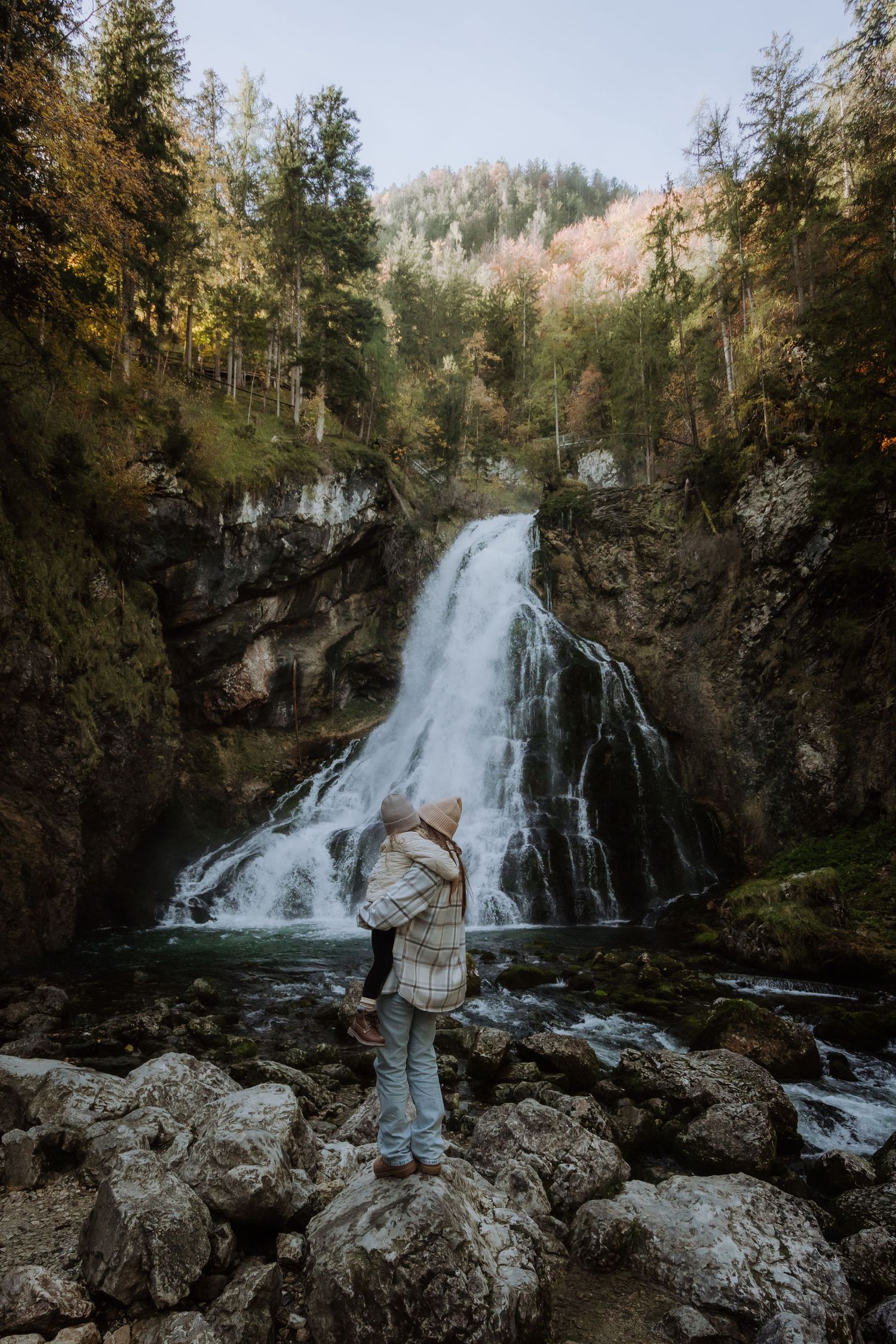 Majestic waterfall cascading down rocky cliffs surrounded by evergreen forest in autumn.
