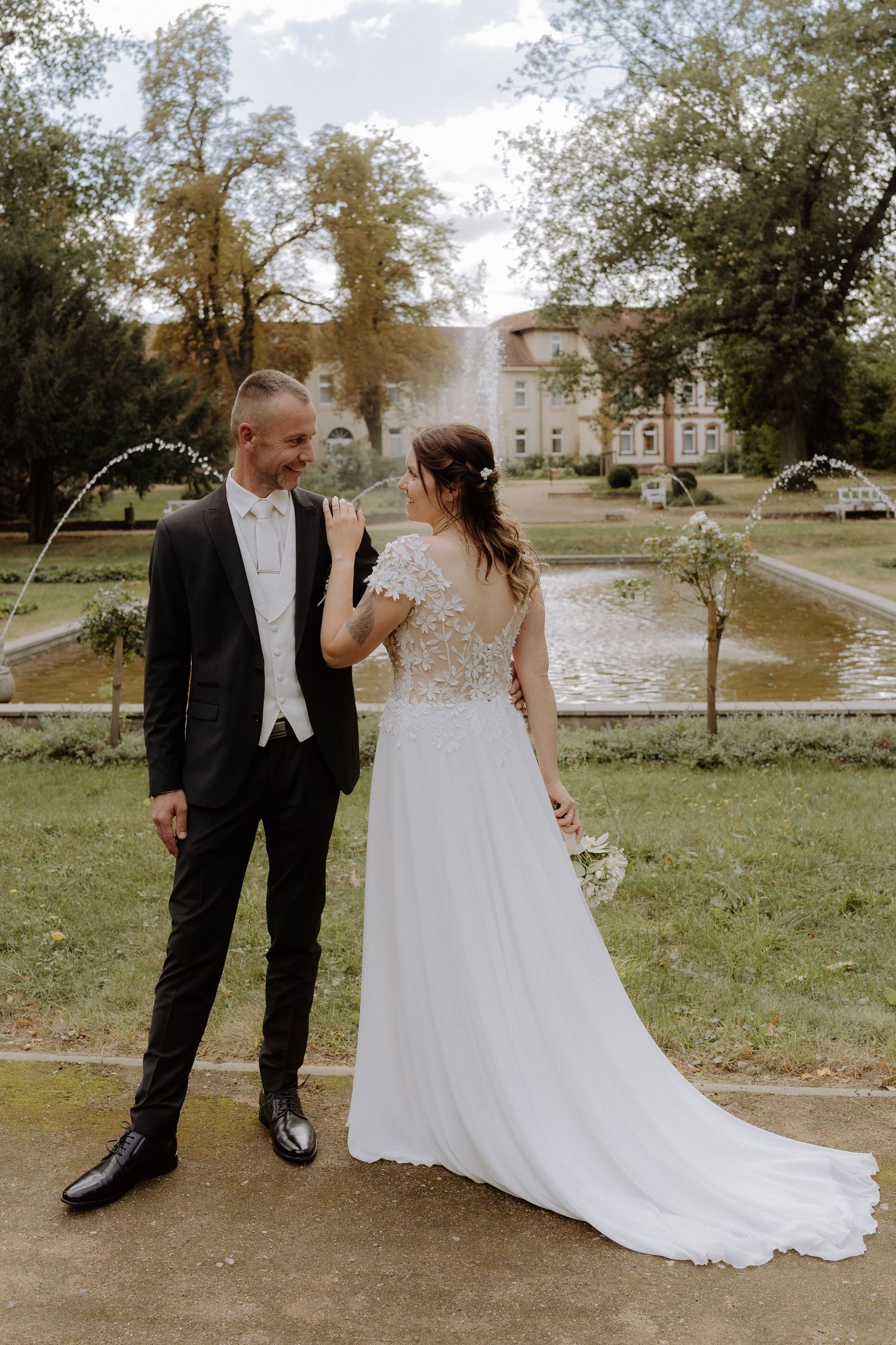 A full-length wedding portrait in a park with fountains and historic building in background.
