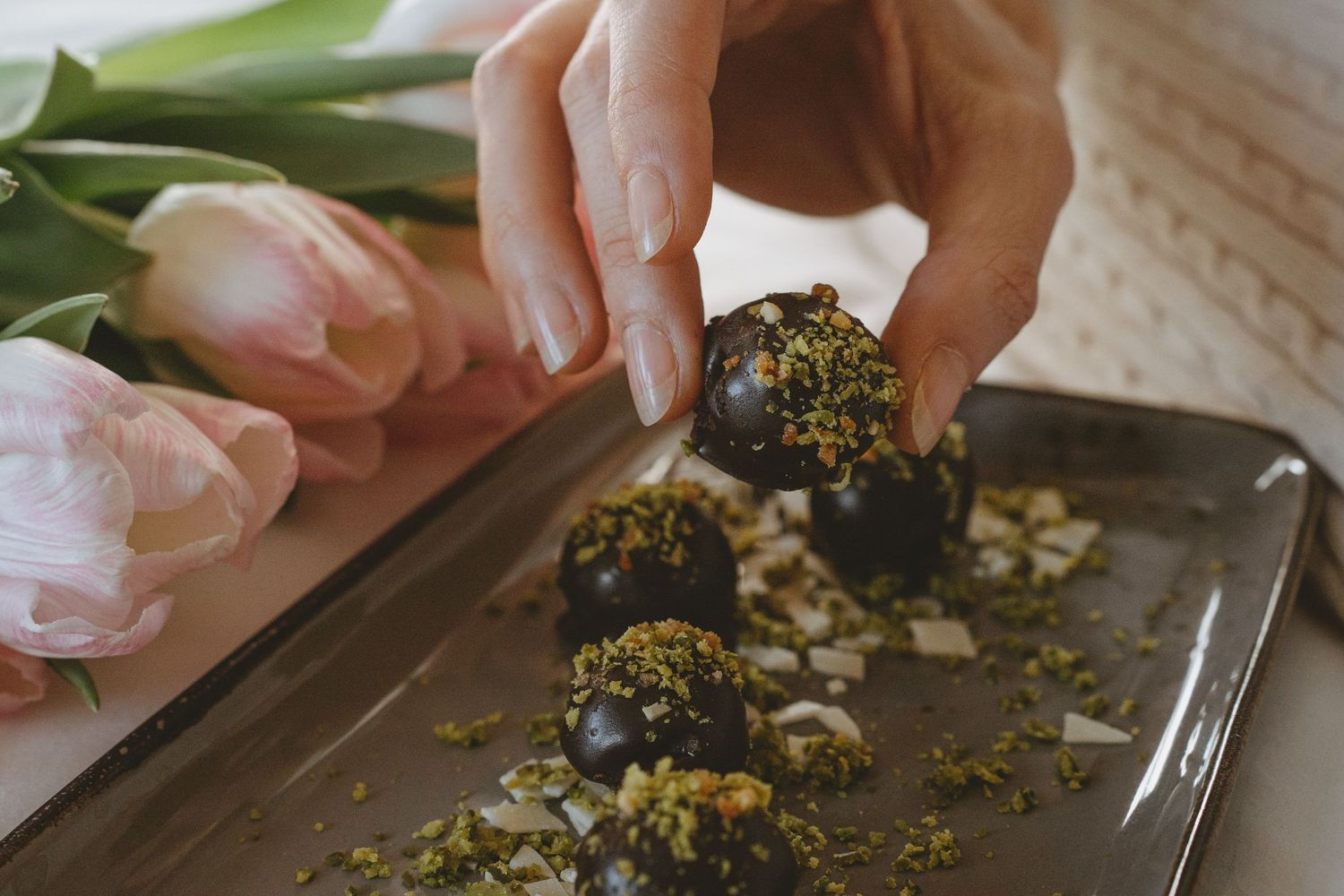 Hands rolling chocolate truffles in chopped nuts on a dark tray with pink tulips.