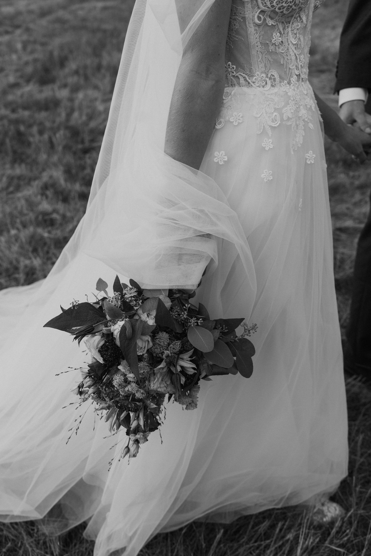 Black and white detail of a flowing wedding dress and bouquet against a grassy field.