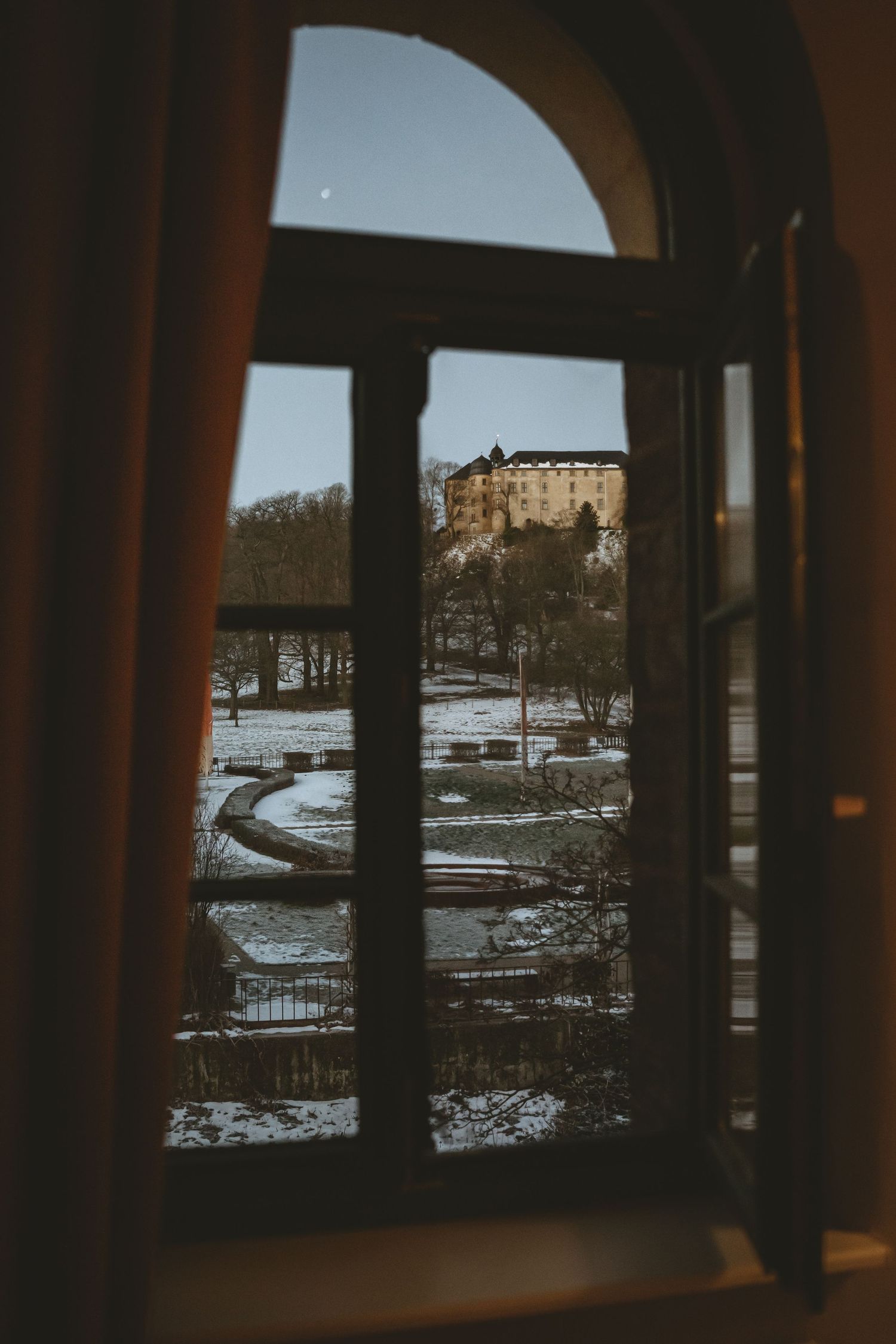 View through arched window of snowy landscape and historic building.