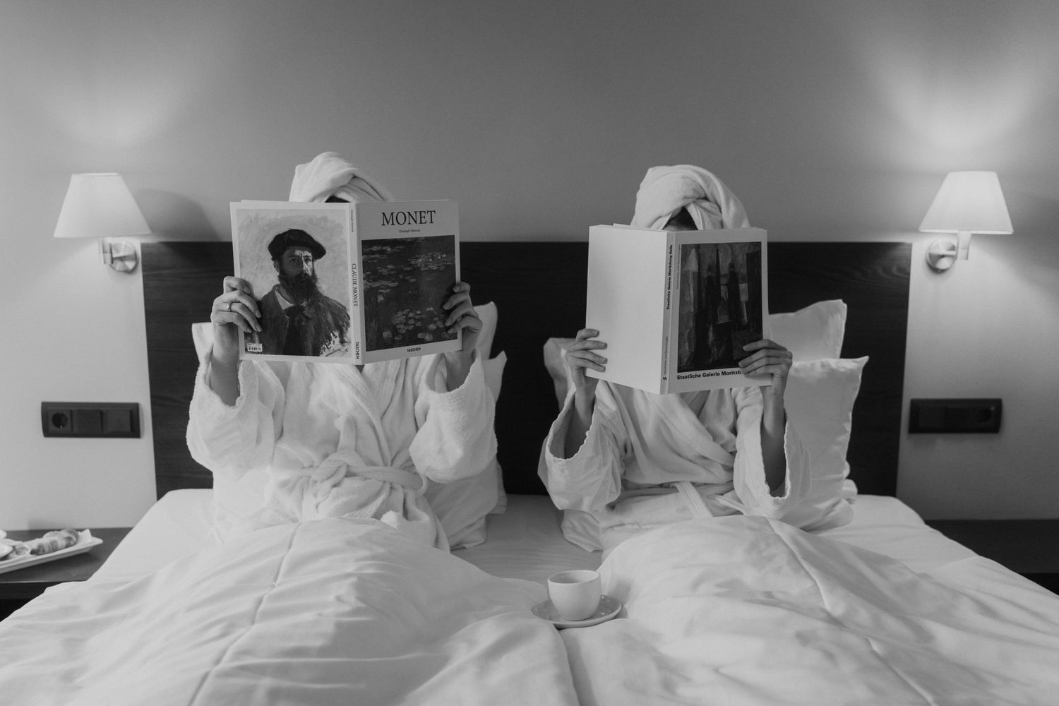 Black and white photo of two people reading newspapers in bed with bedside lamps.