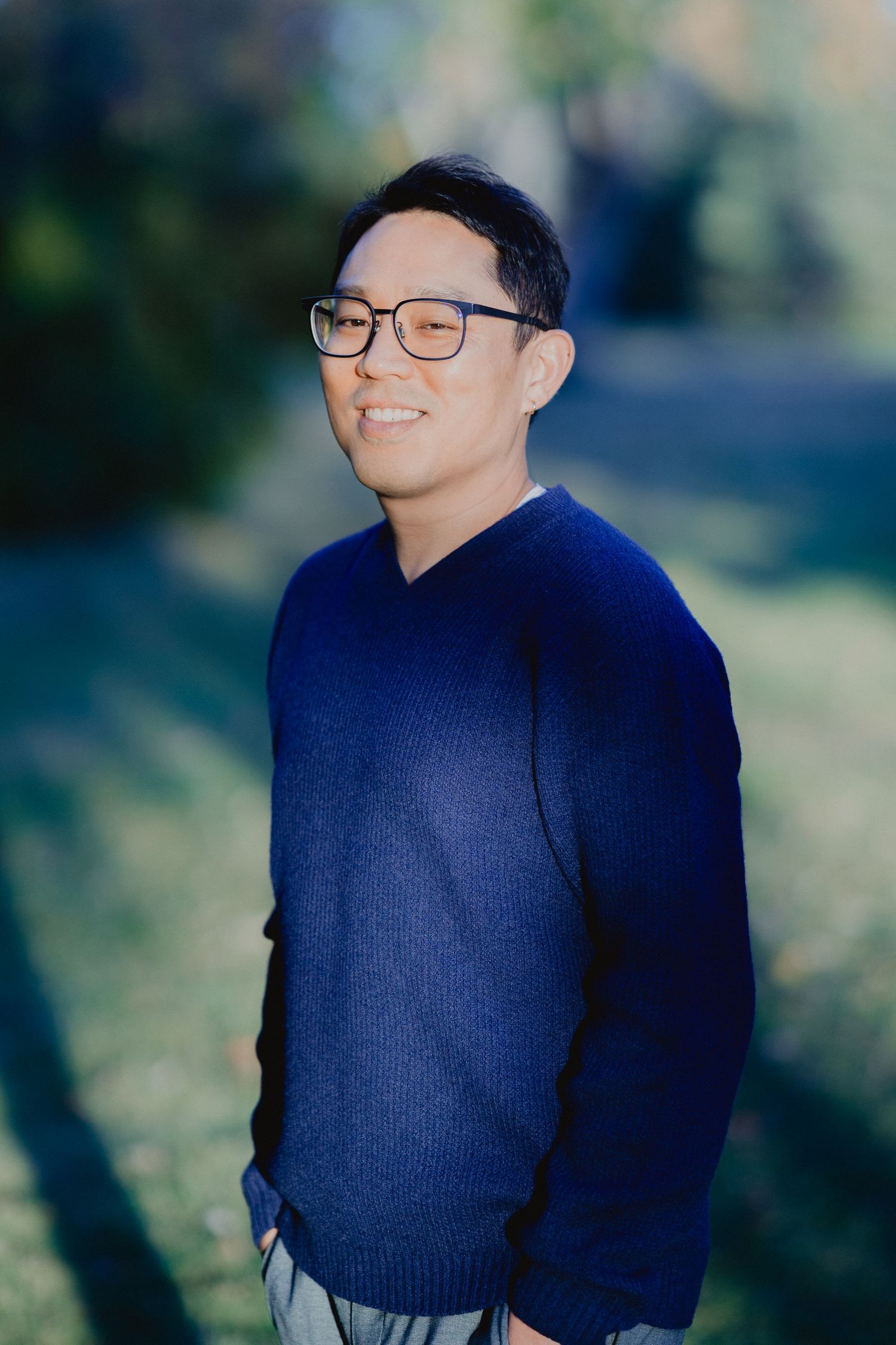 A person wearing a navy blue sweater poses outdoors against a blurred natural background in a photo series.