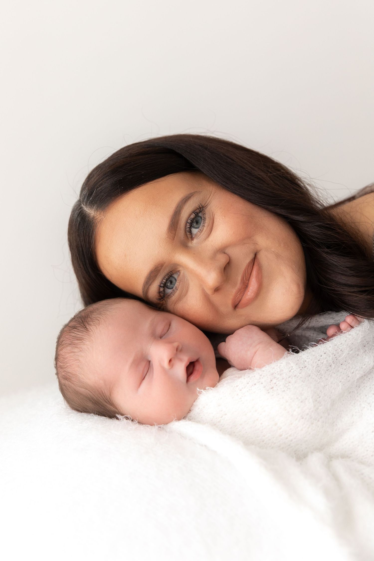 A sleeping newborn baby swaddled in white blanket lies next to a smiling parent on white background.