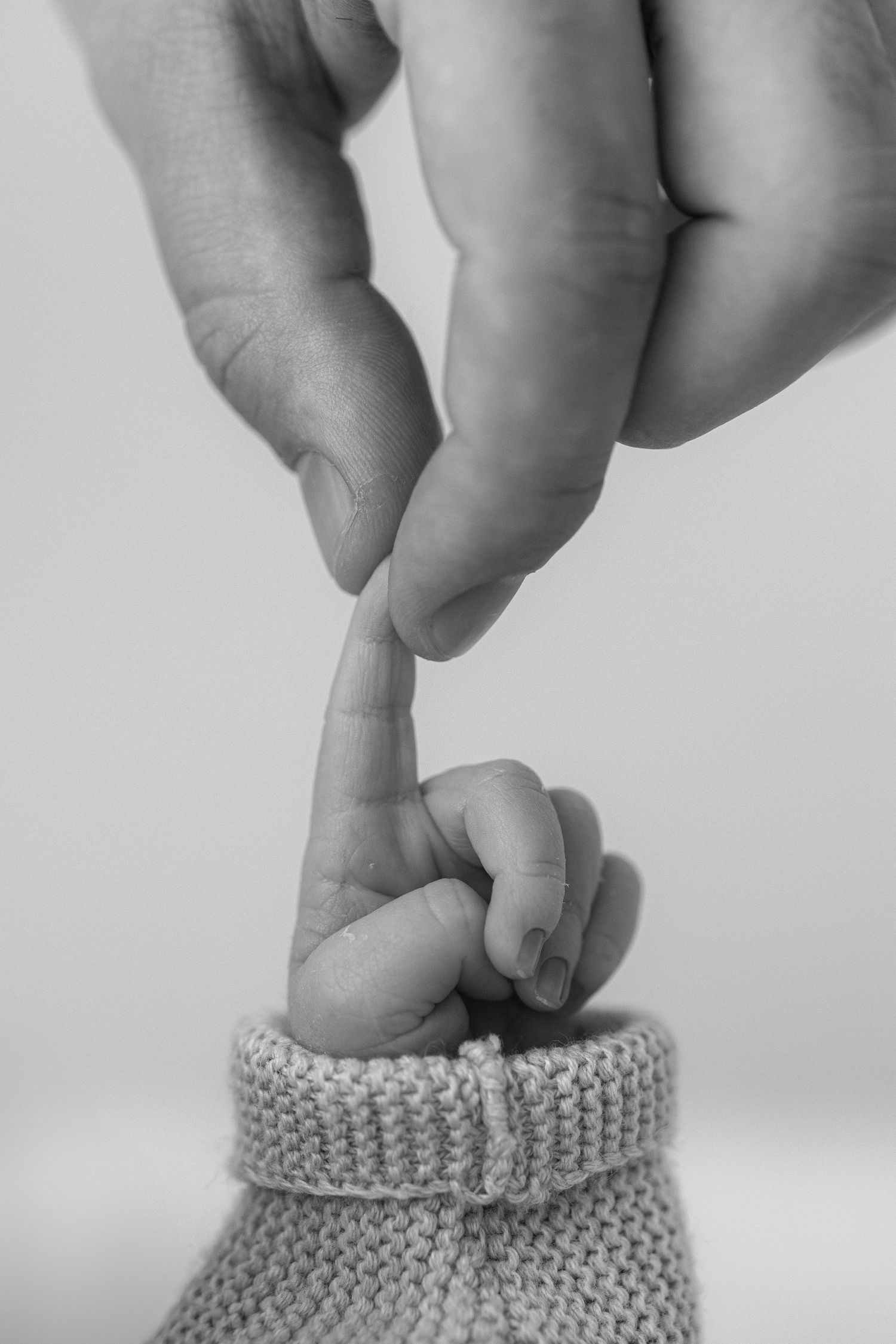 A tender moment as an adult's finger gently touches a baby's tiny foot in a knitted grey bootie, captured in black and white.
