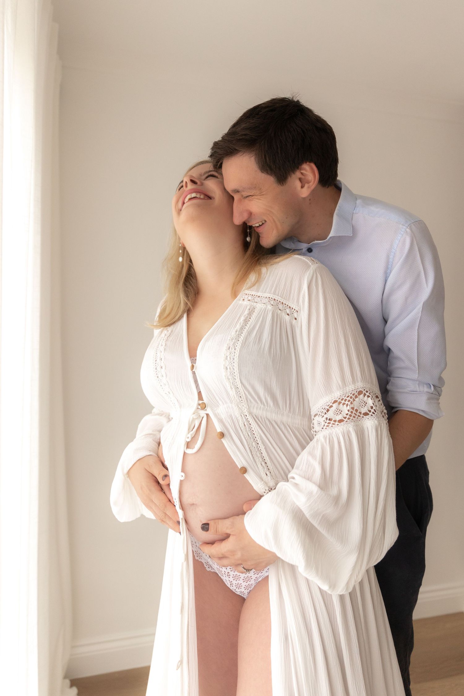 Intimate maternity portrait in white lace dress against light wall.