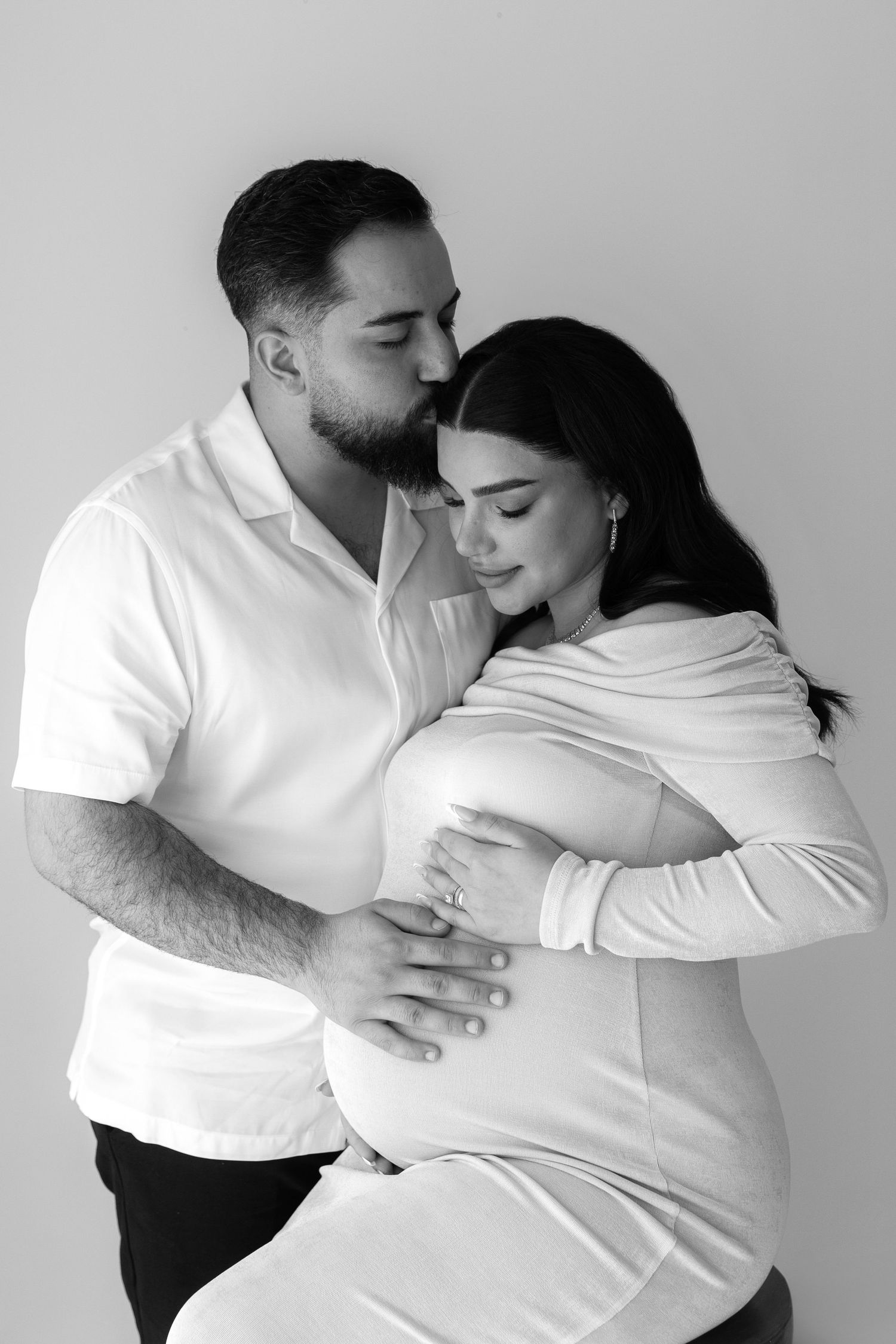 Black and white maternity photo of expectant parents sharing an intimate moment in matching white outfits.