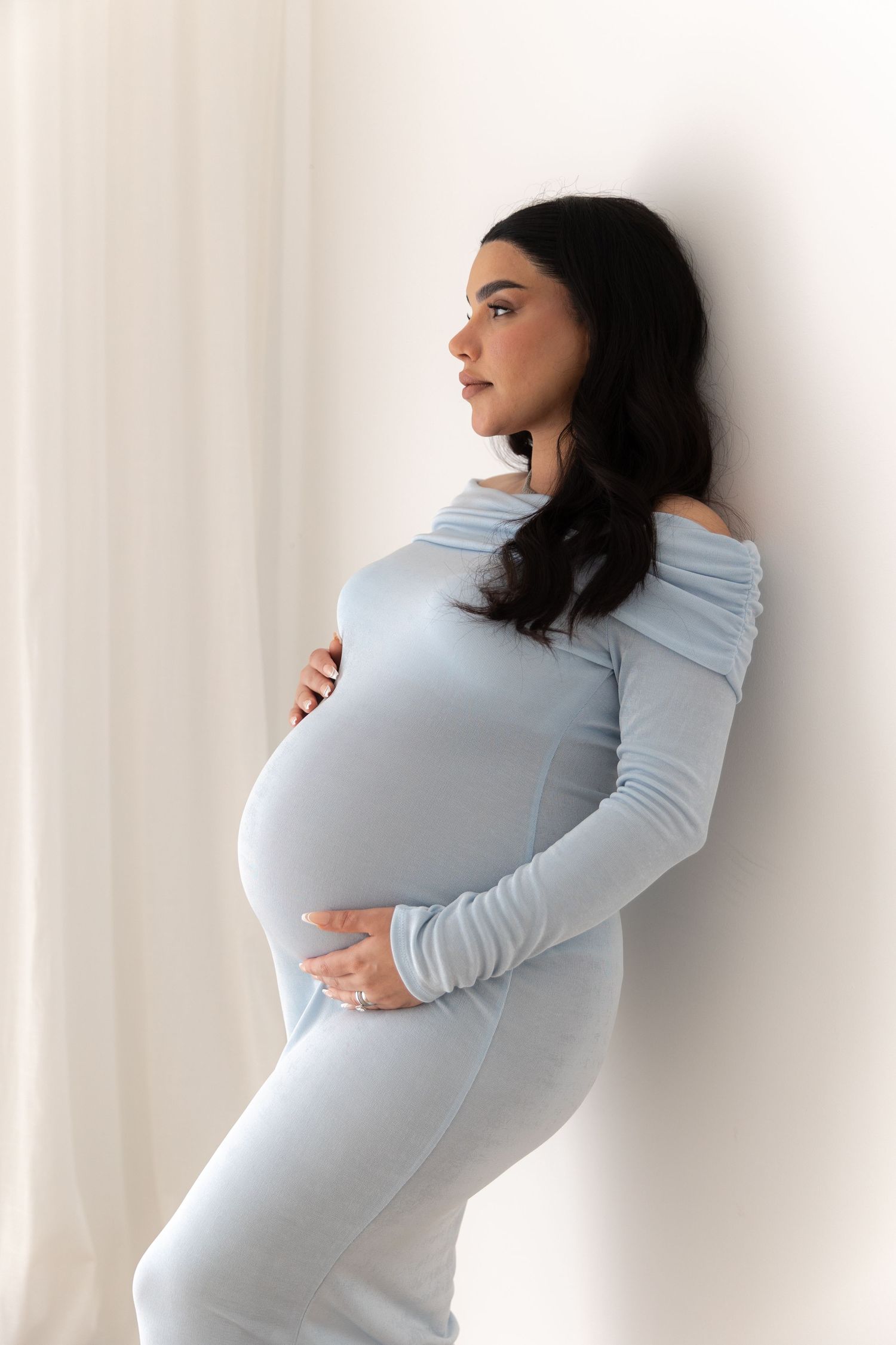 Expecting mother in pale blue fitted dress poses against white curtain background highlighting her pregnancy.