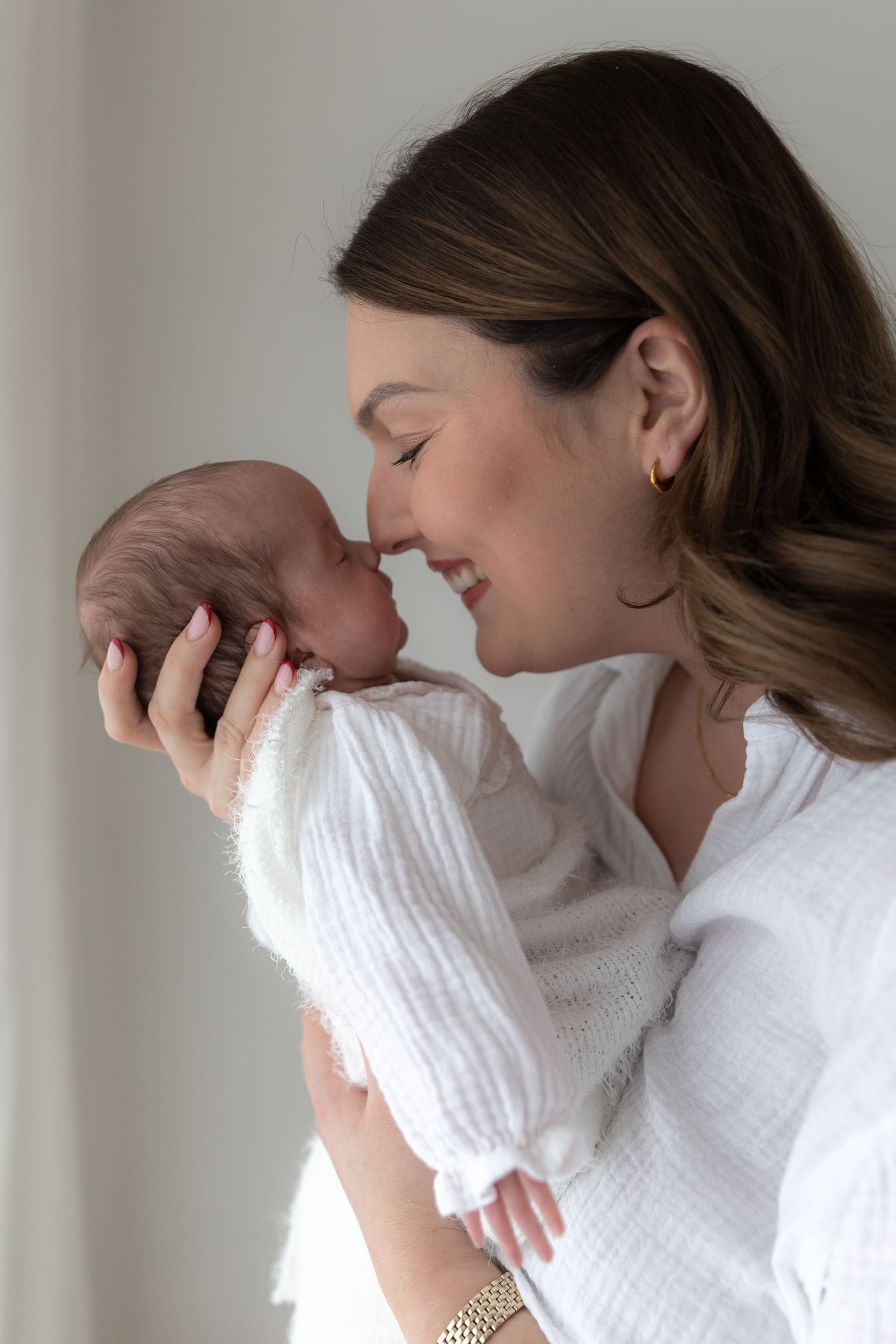 Intimate moment between mother and newborn in white attire against neutral background.