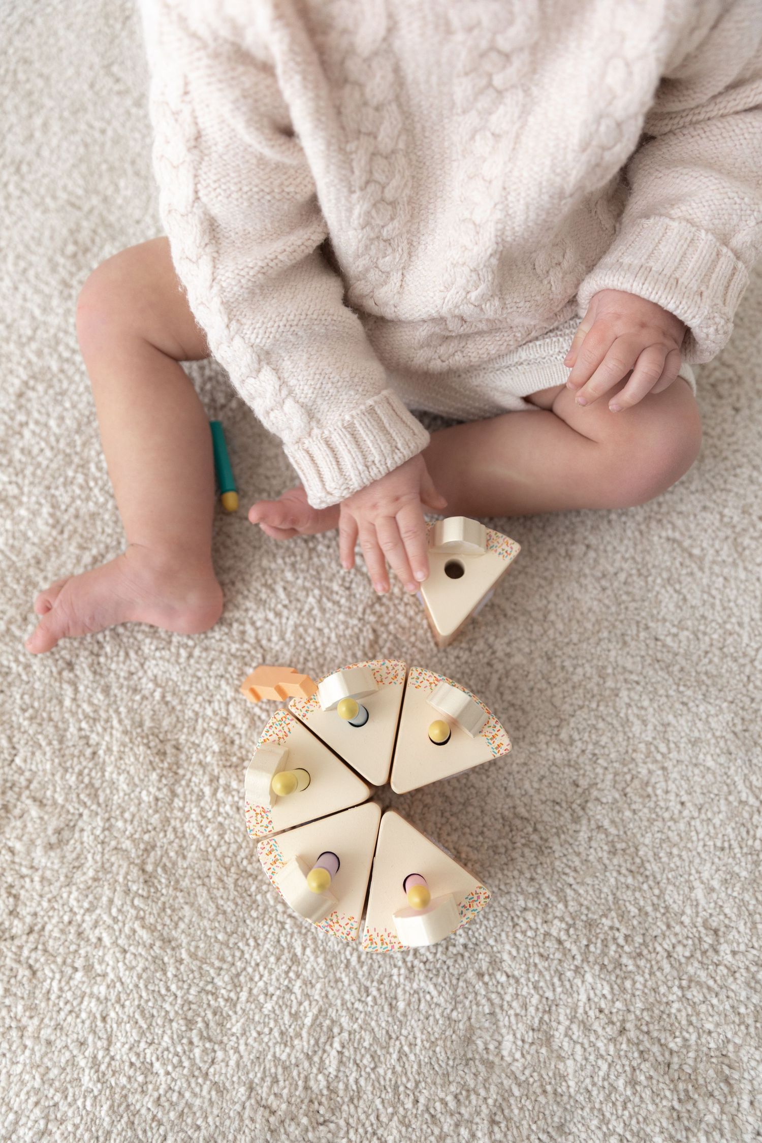 Baby in cream sweater playing with wooden toy pieces on carpet.