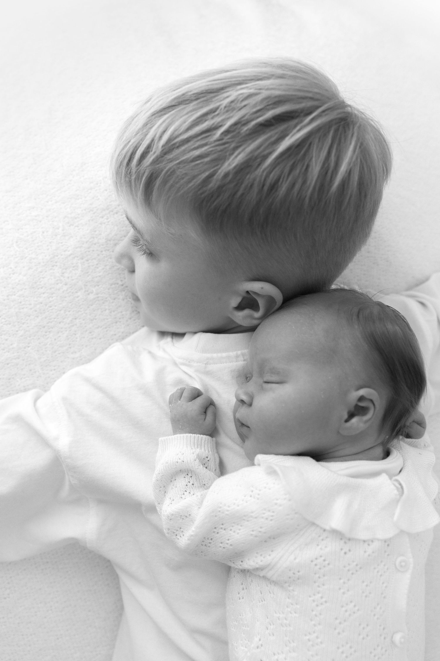 Two children resting together on white bedding in tender black and white portrait.
