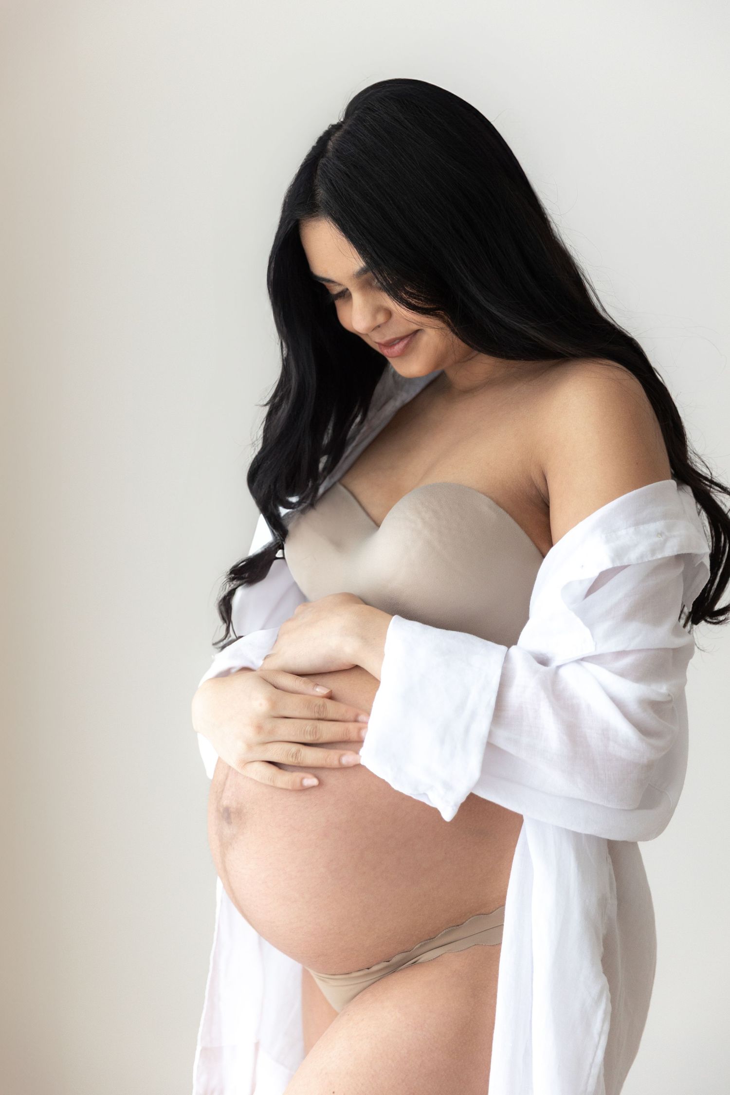A tender maternity portrait of an expectant mother in a white open shirt and nude bra cradling her baby bump.