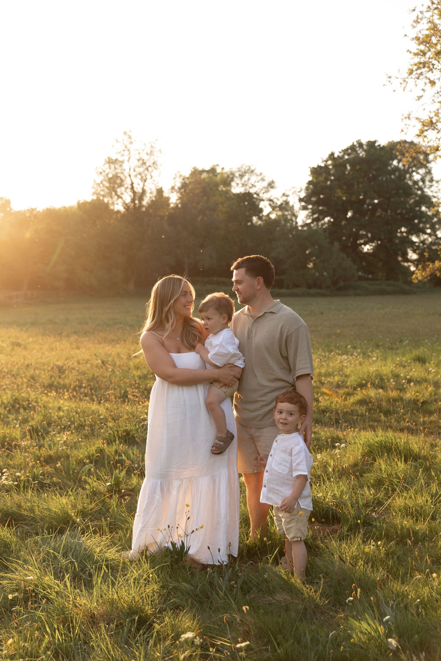 Silhouetted family stands together in a sunlit grassy field at golden hour with trees in the background.